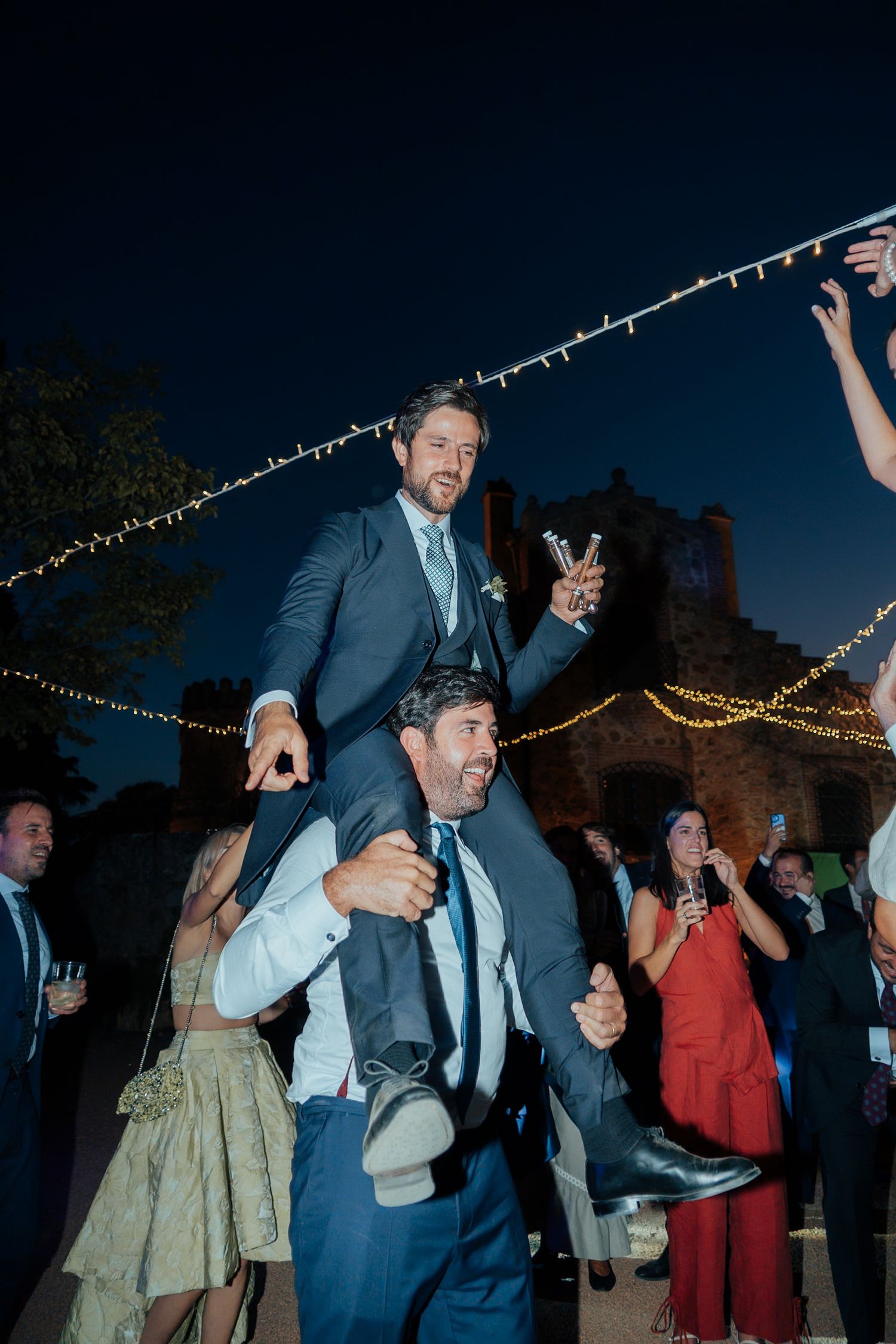 Excited wedding guest lifted on shoulders during lively outdoor evening wedding celebration.