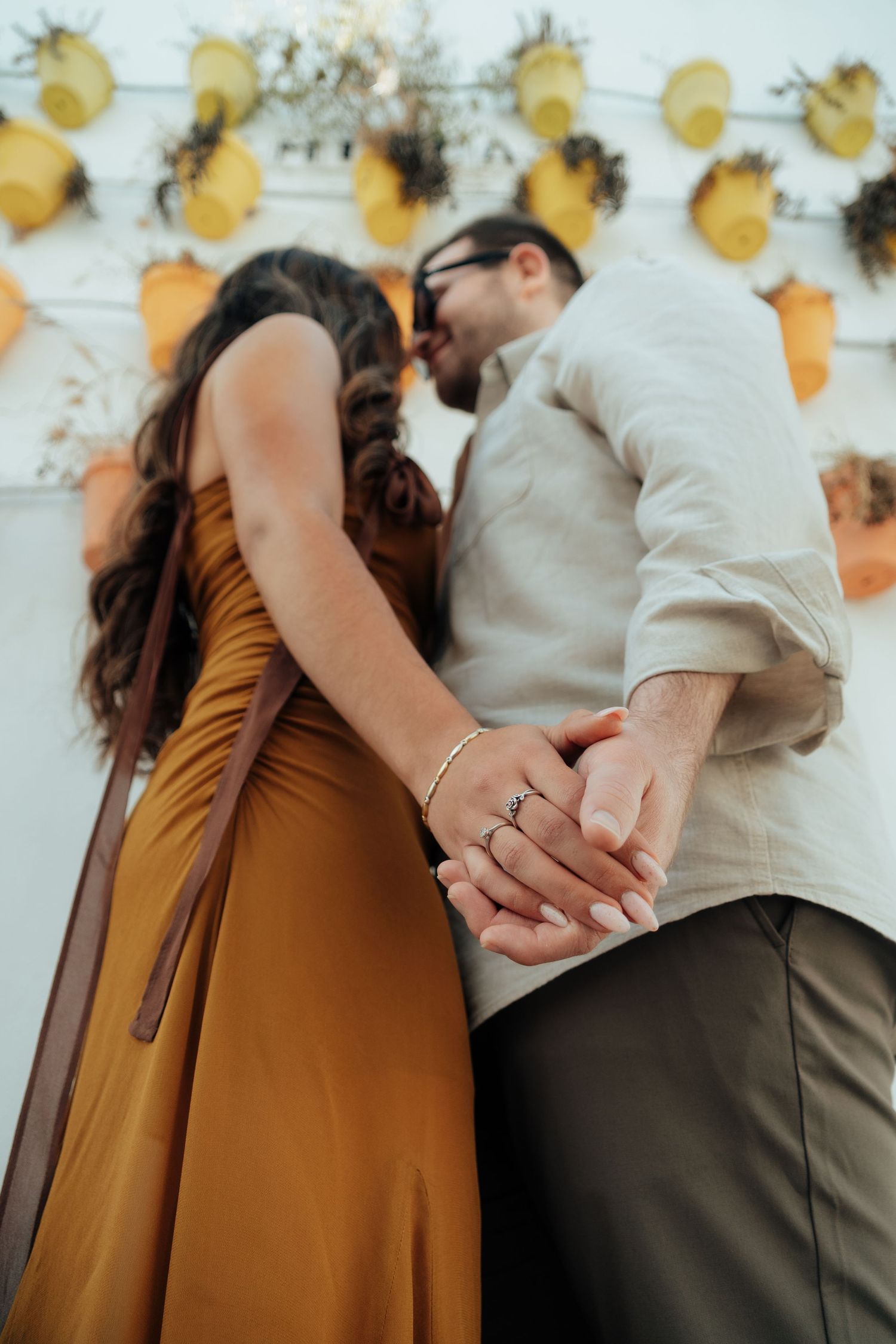 Couple holding hands and kissing in front of a white wall decorated with yellow flowers and plants.