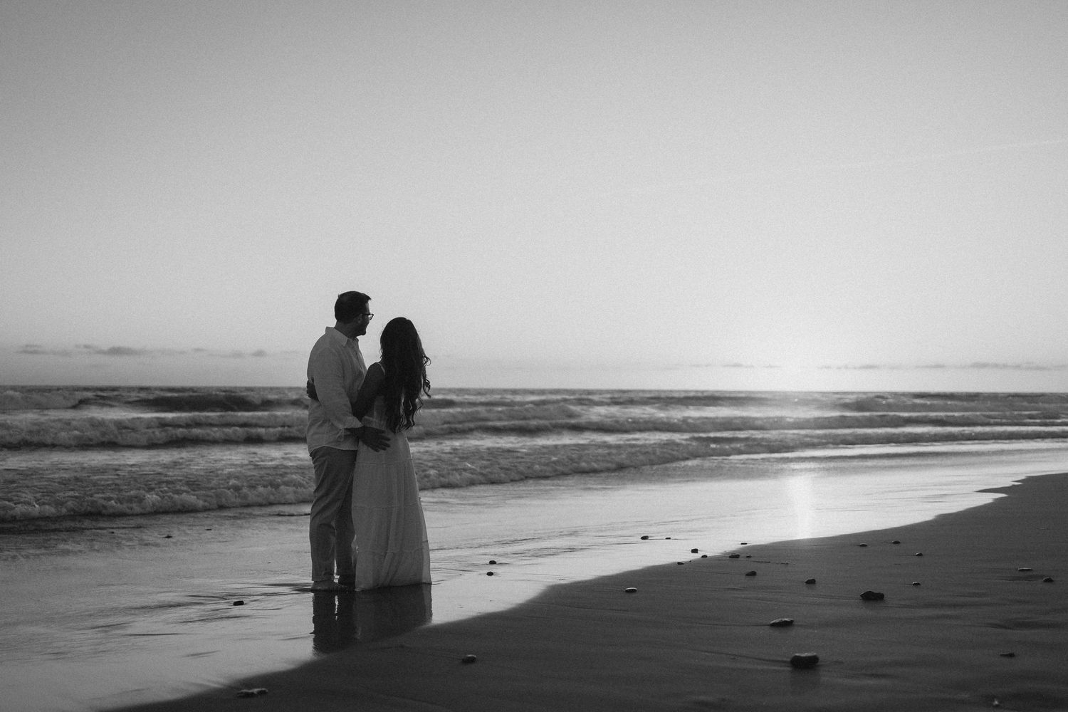 Black and white photo of a couple standing together on a beach at sunset, waves gently lapping the shore.