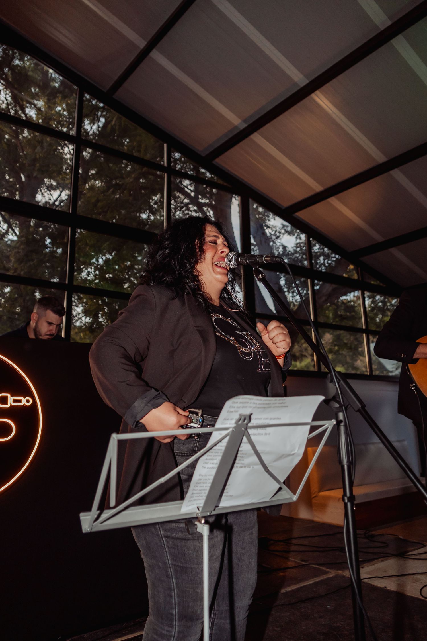 Woman singing passionately at a microphone stand with sheet music during a live indoor performance.
