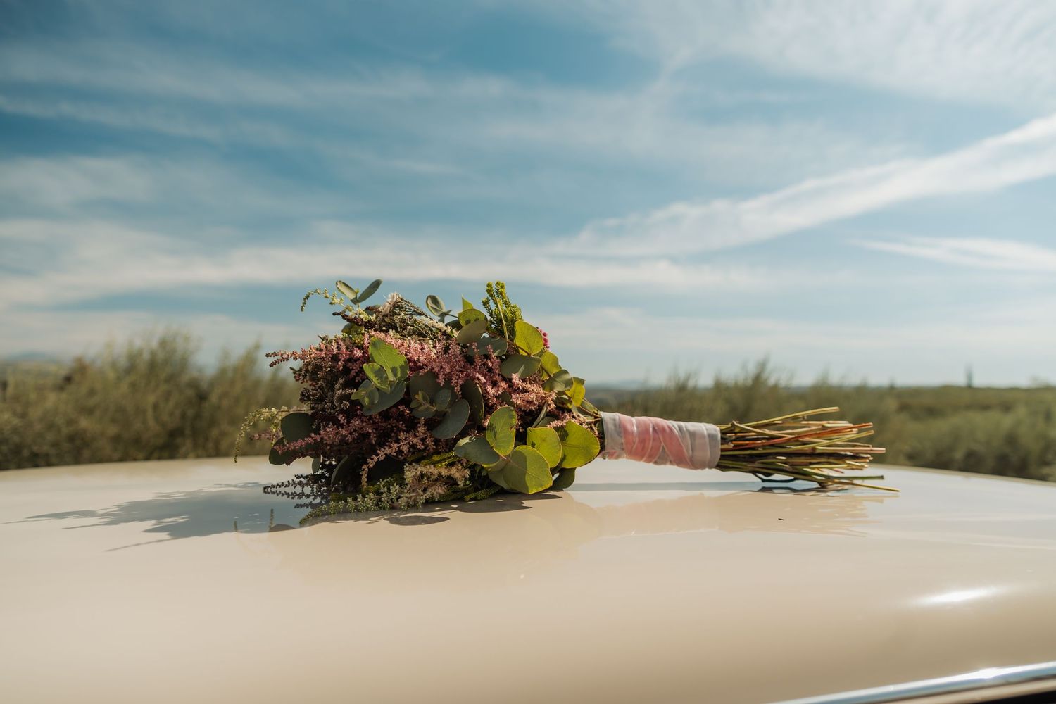 Rustic wildflower bridal bouquet with greenery resting on a vintage cream car hood outdoors.