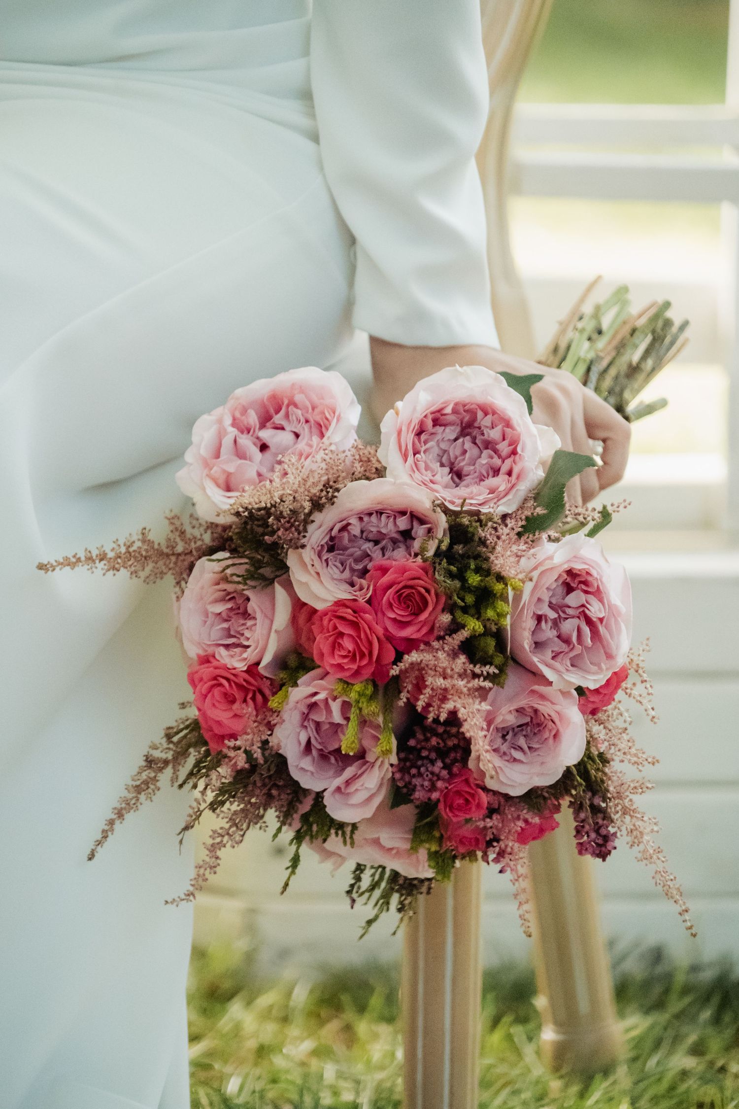 Bride holding a lush pink peony and rose wedding bouquet beside a white chair outdoors.