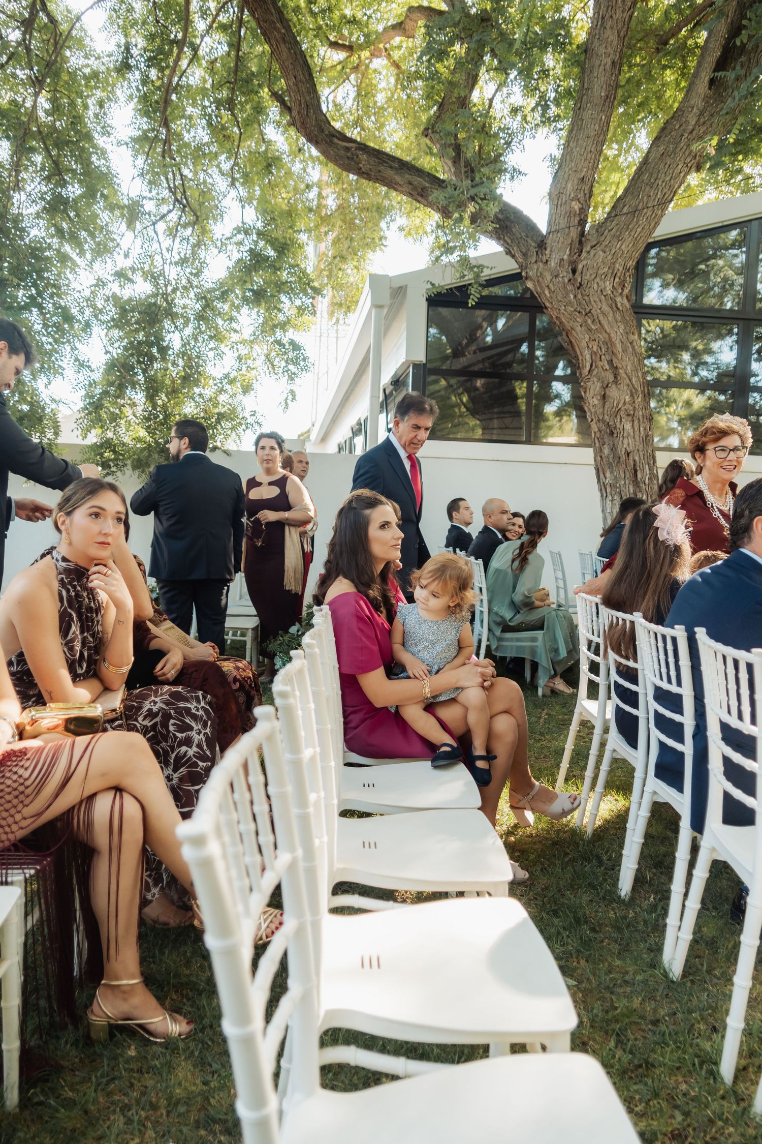 Wedding guests seated in white chairs on grass await outdoor ceremony beneath large shady trees.