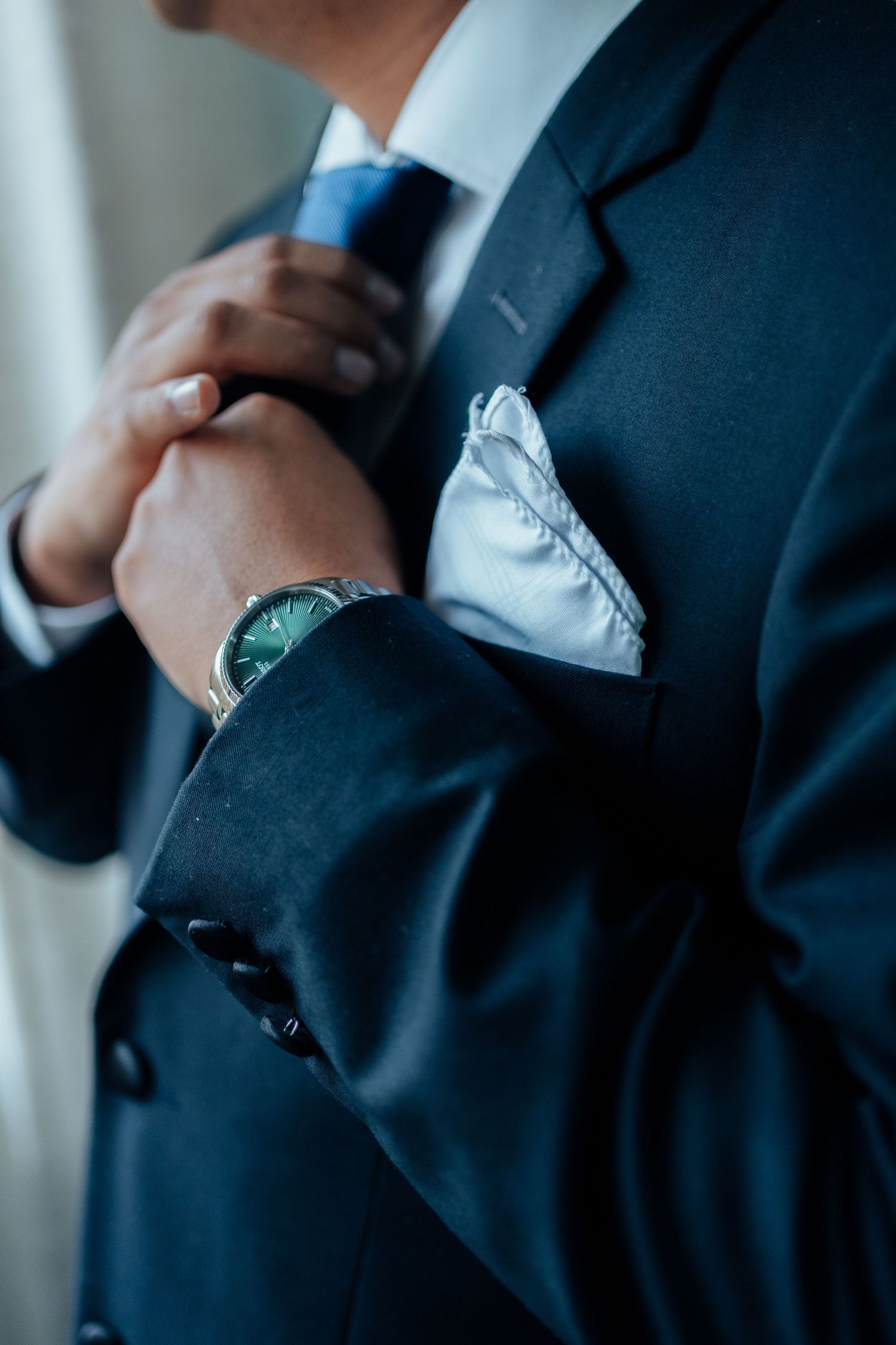 Close-up of a groom adjusting his blue tie wearing a navy suit with white pocket square and watch.