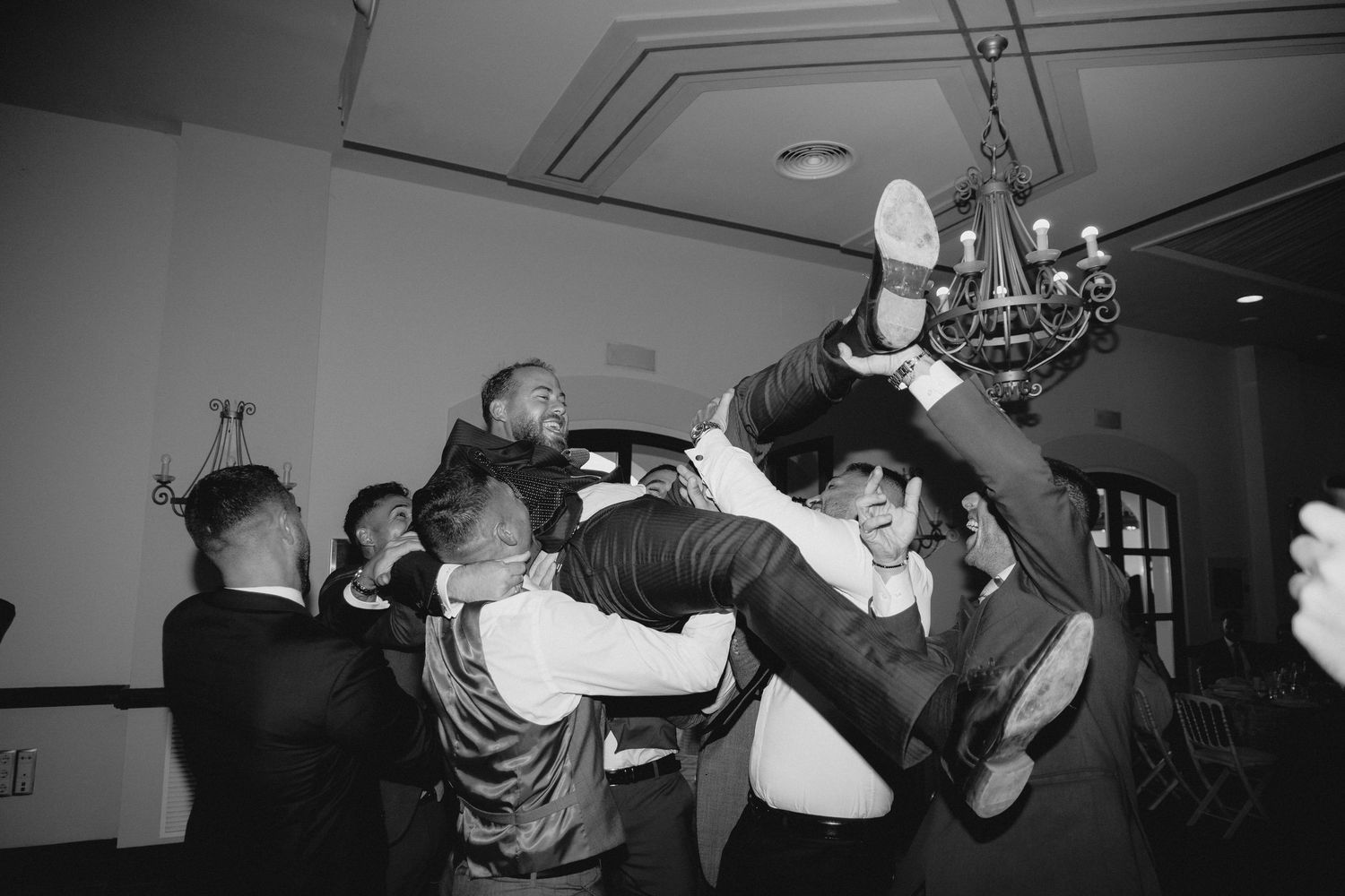 Groomsmen lifting groom in the air during lively wedding reception celebration in black and white.