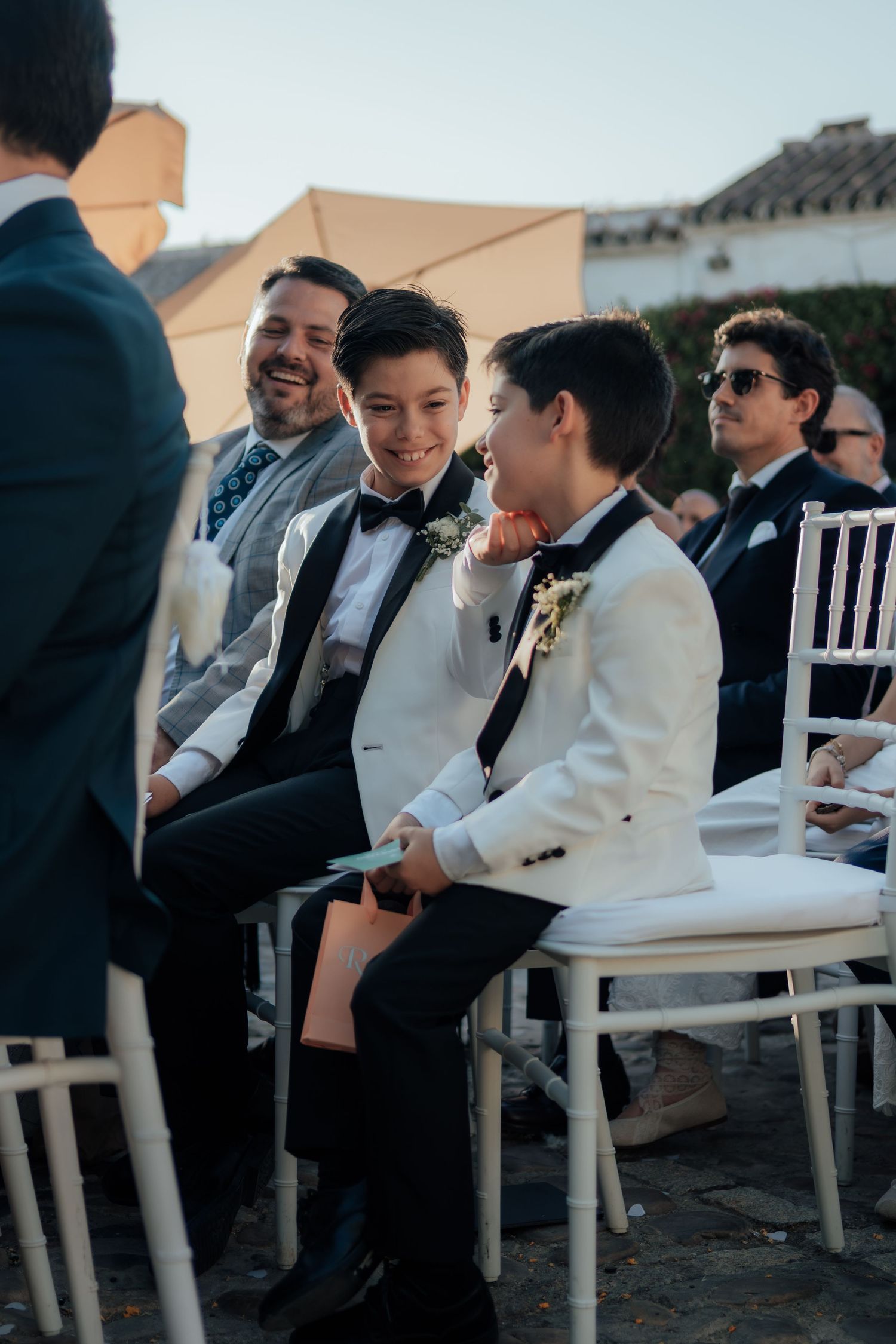 Two young boys in white tuxedos laugh together while seated at outdoor wedding ceremony.