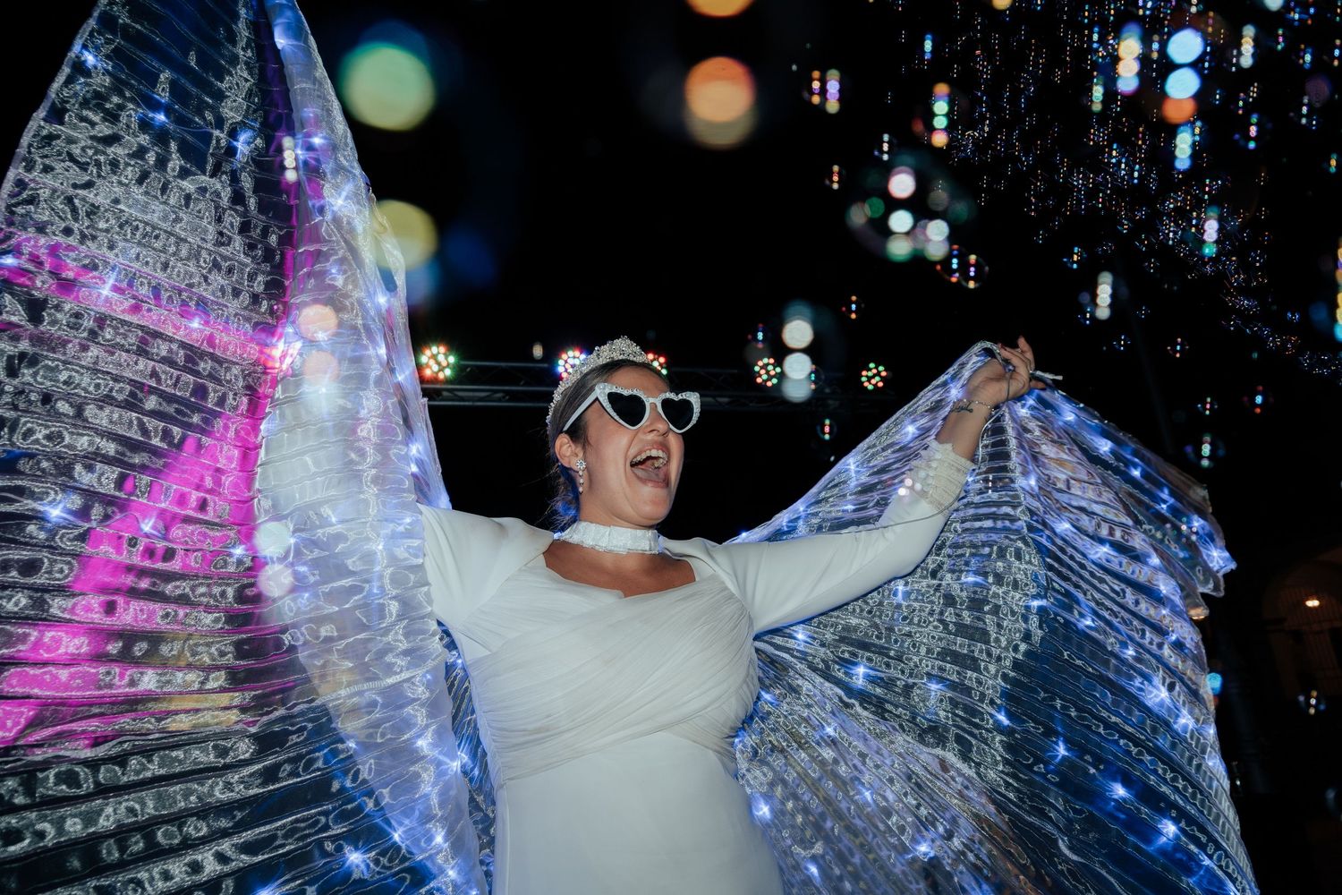 A joyful bride in white with heart sunglasses spreads glowing LED butterfly wings outdoors at night.