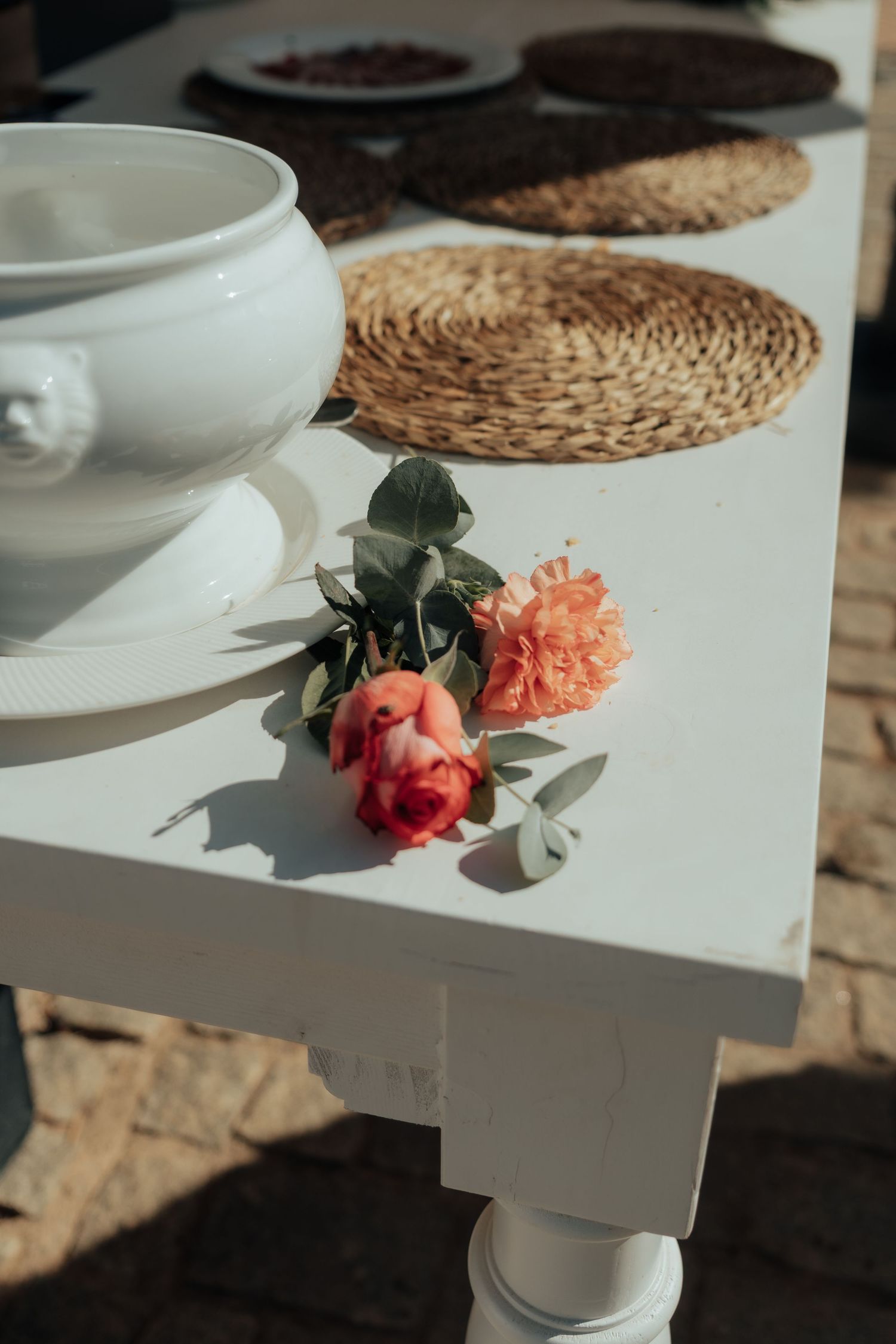 Two pink roses rest on a white table beside a ceramic bowl and woven placemats.