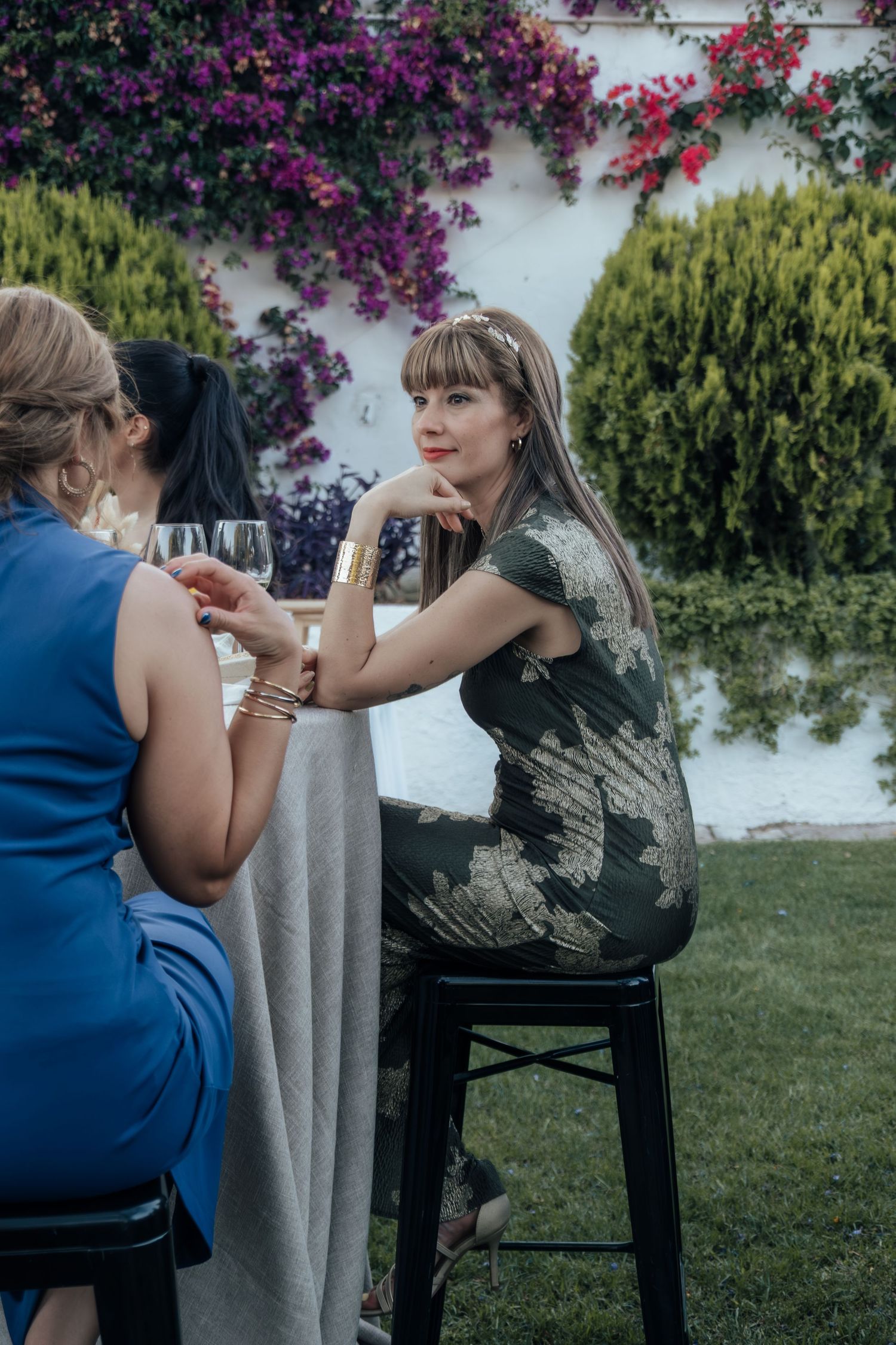 Elegant wedding guest in floral dress sitting at outdoor cocktail table with wine glass.