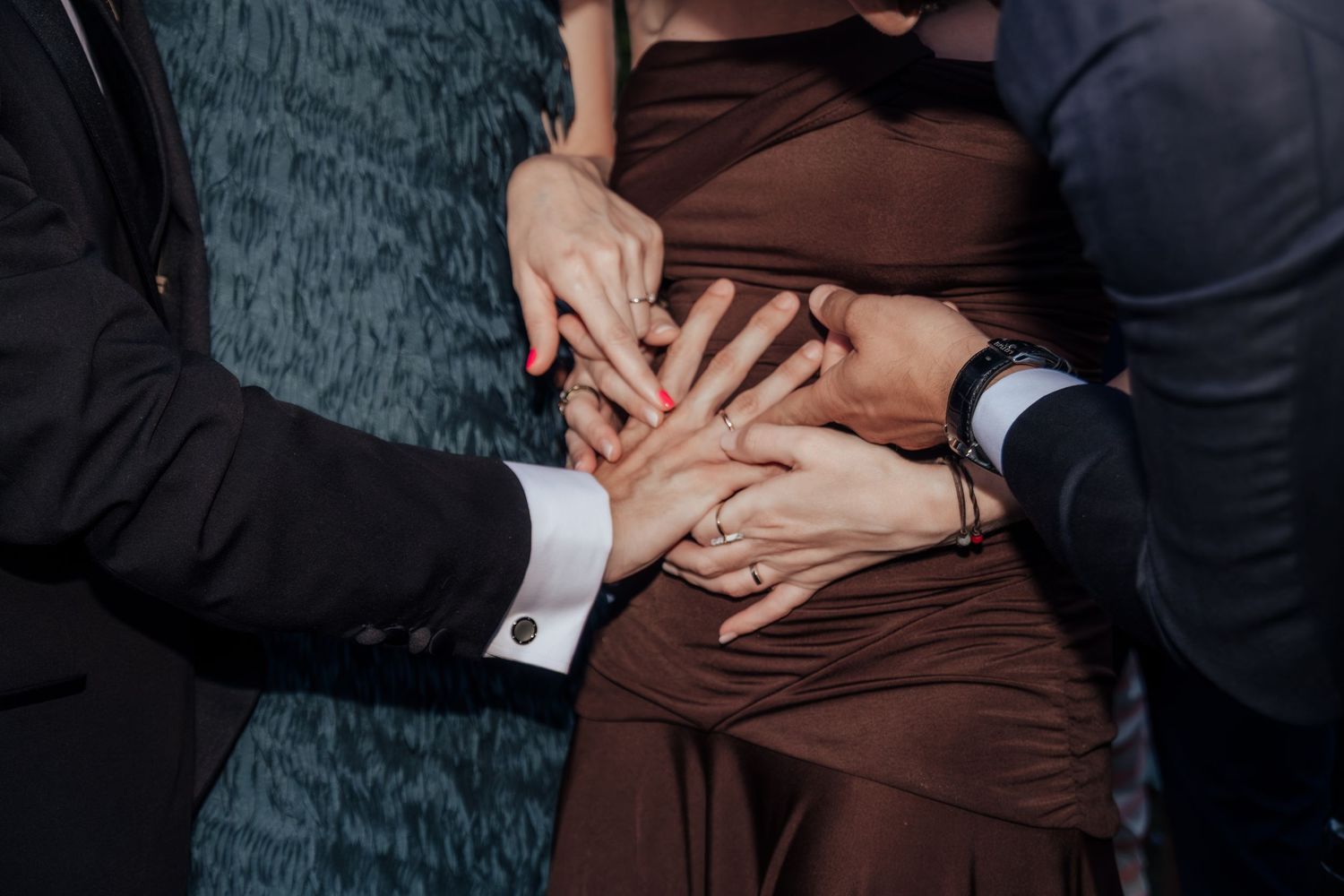 Close-up of multiple hands touching a pregnant woman's belly in a brown dress at a celebration.