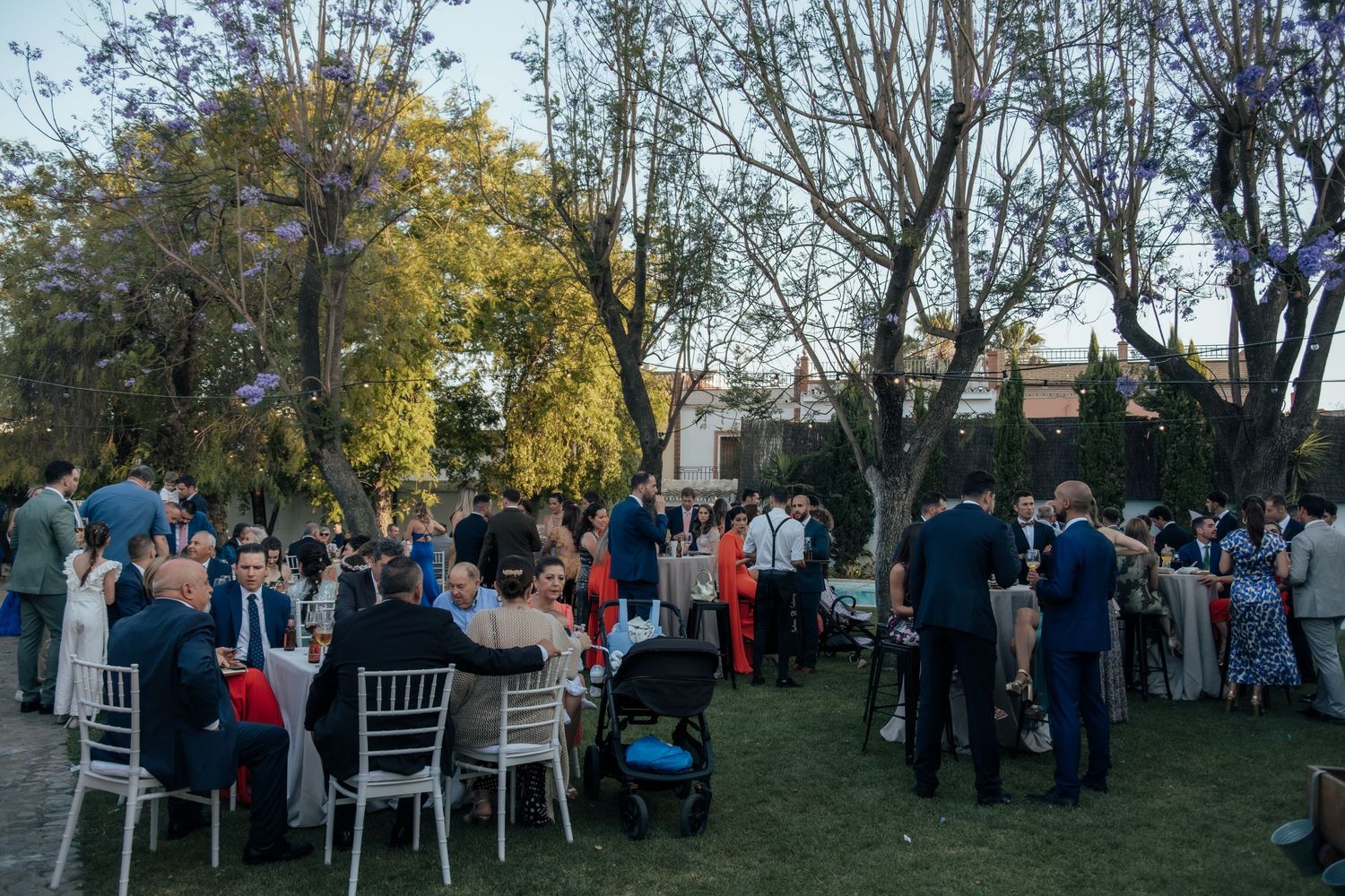 Outdoor wedding reception on a green lawn with guests seated at white chairs around decorated tables under trees.