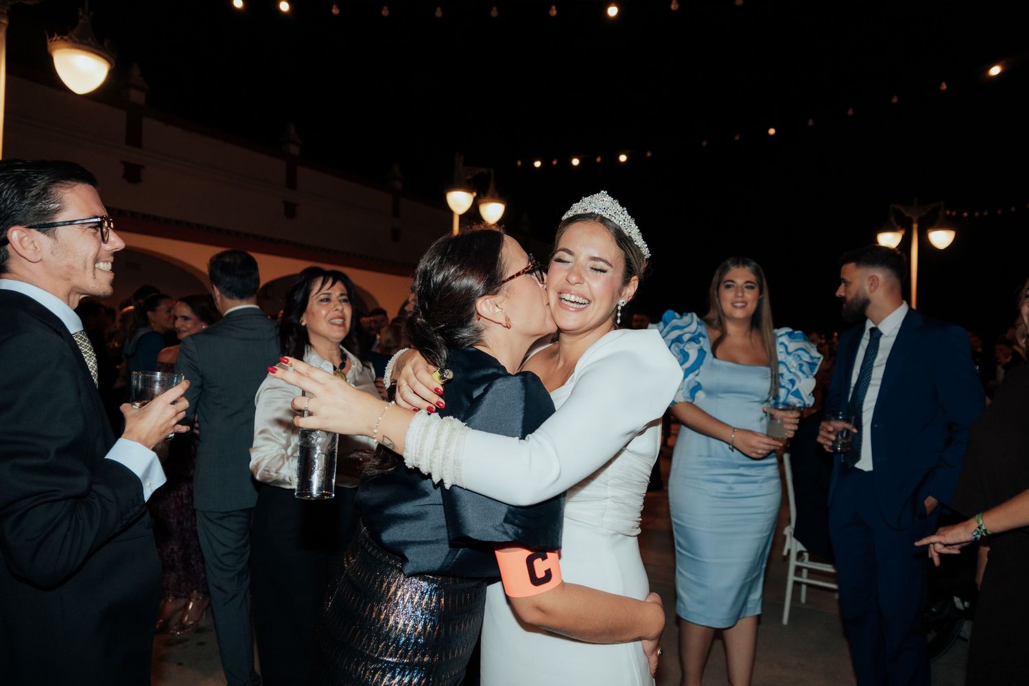 Bride wearing tiara embracing guest warmly at lively indoor wedding reception celebration.