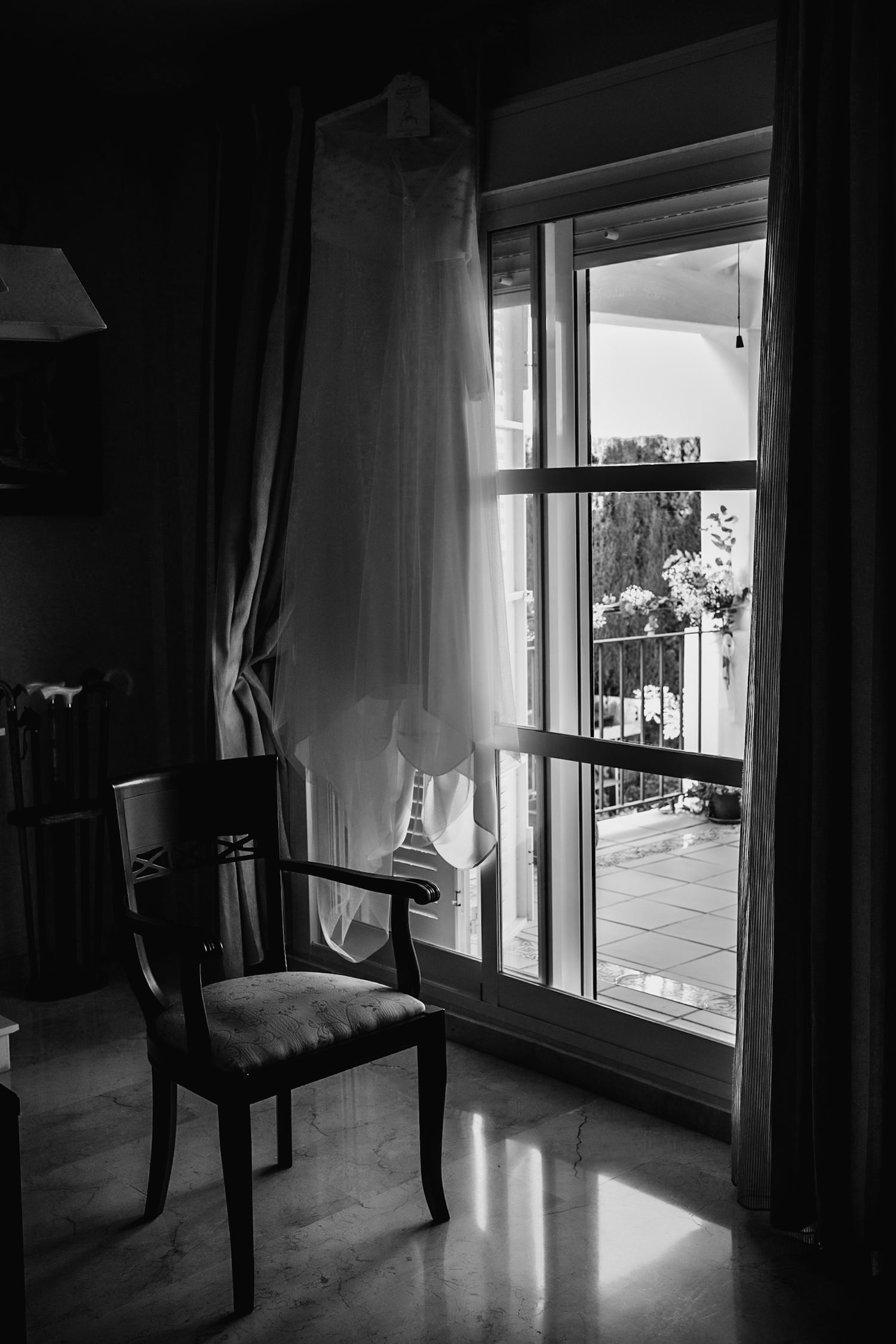 Black and white image of a wedding dress hanging by a sunlit balcony door beside a wooden chair.