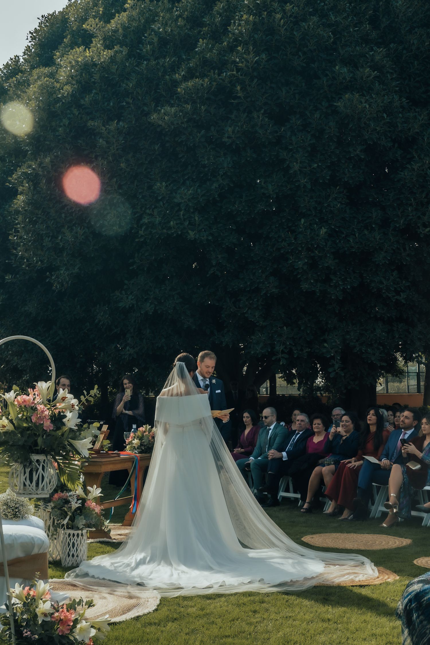 Bride in a long white veil standing at the altar during an outdoor wedding ceremony with seated guests.