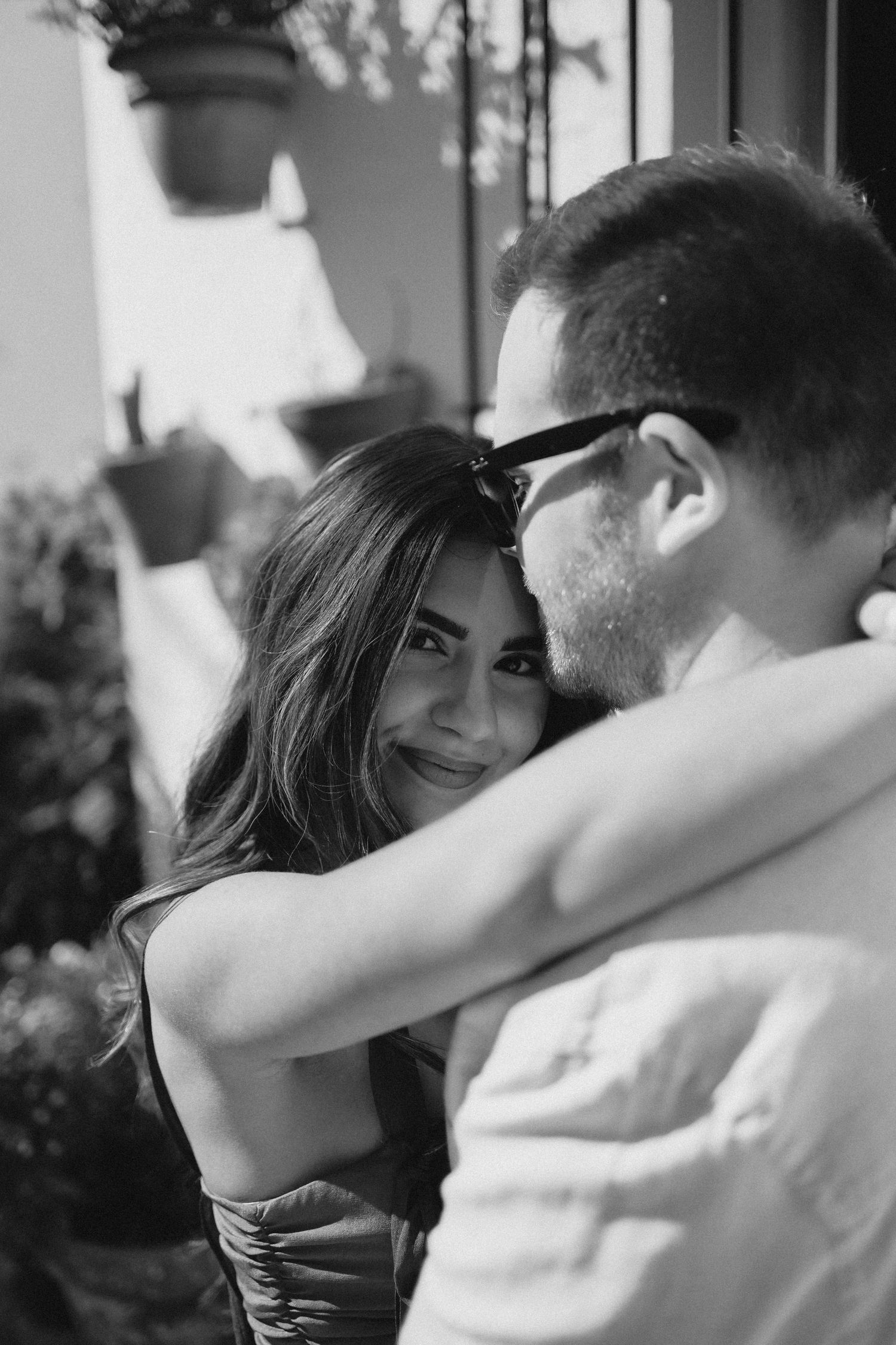 Black and white photo of a smiling woman hugging a man wearing glasses outdoors near potted plants.