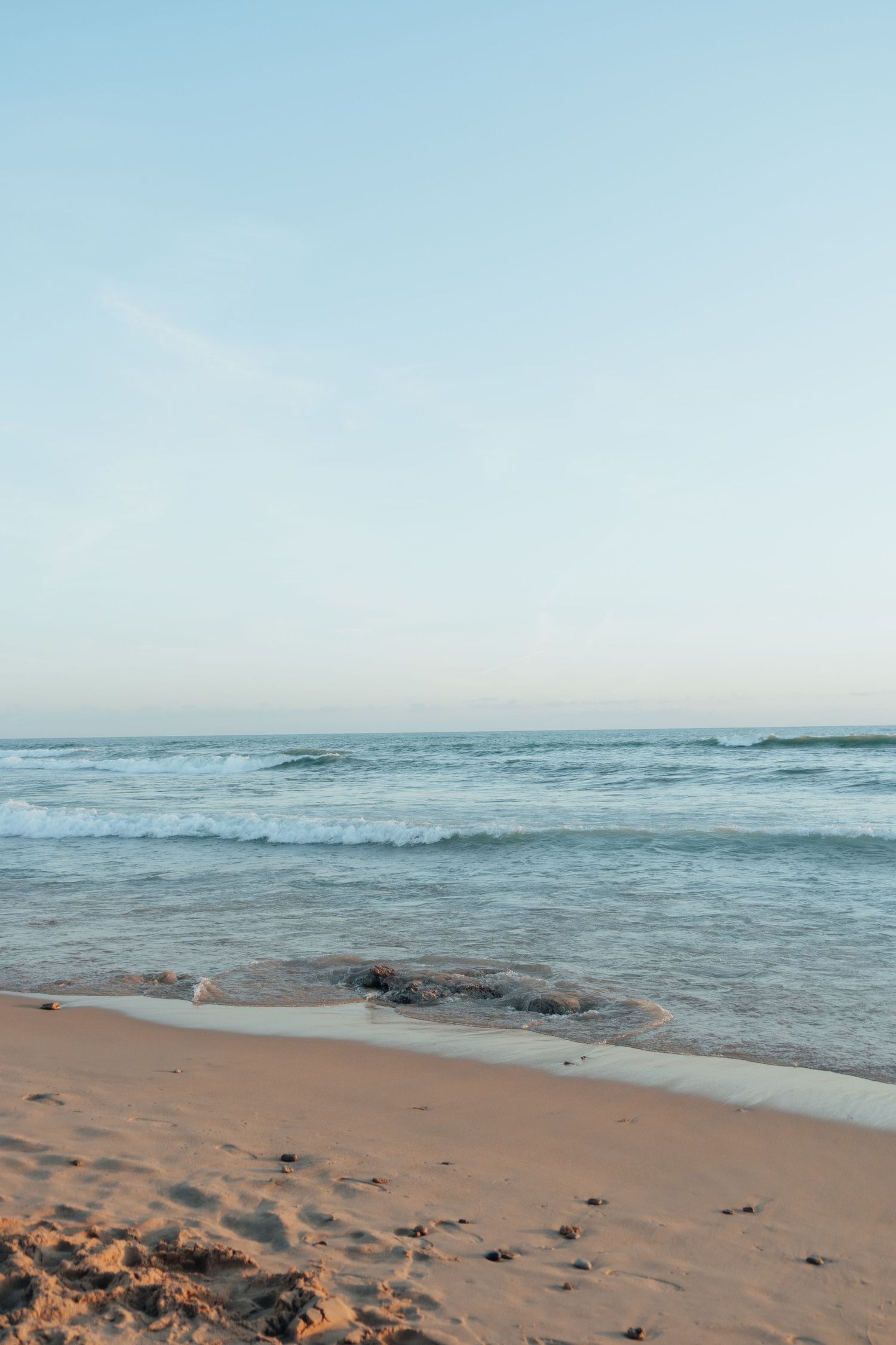 Calm ocean waves gently washing onto a sandy beach under a clear pale blue sky at dusk.