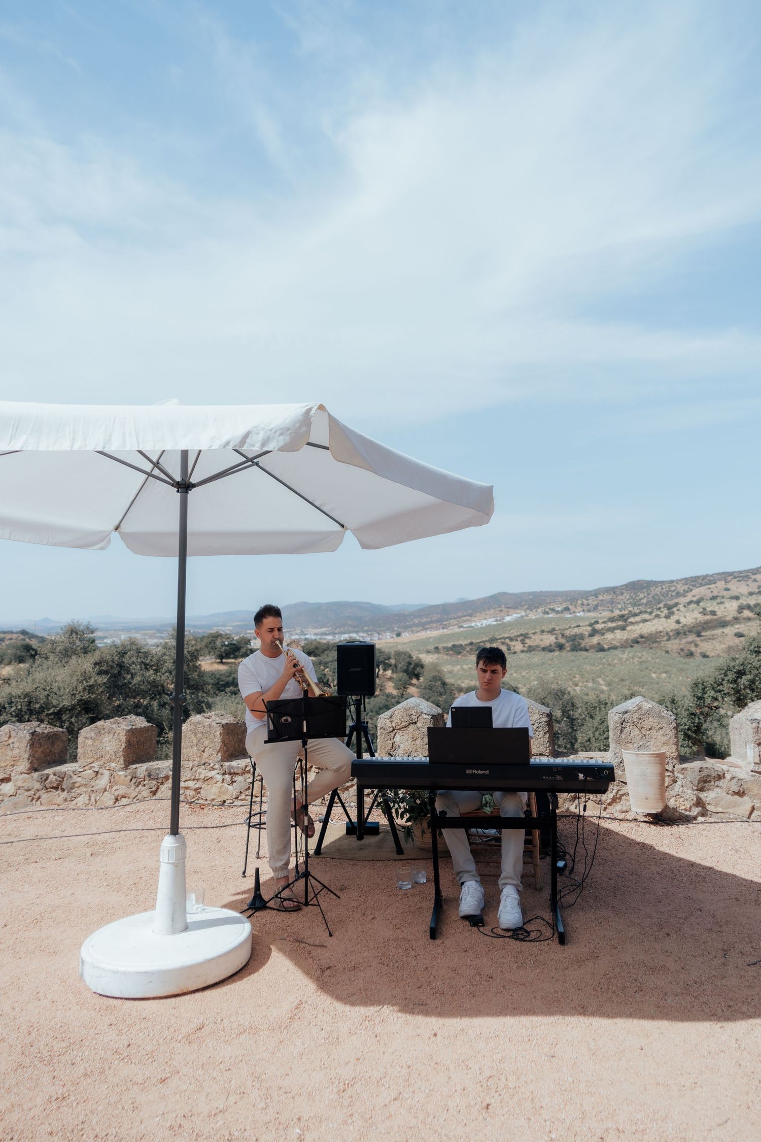 Two musicians performing with keyboard and trumpet under white umbrella at scenic outdoor wedding.
