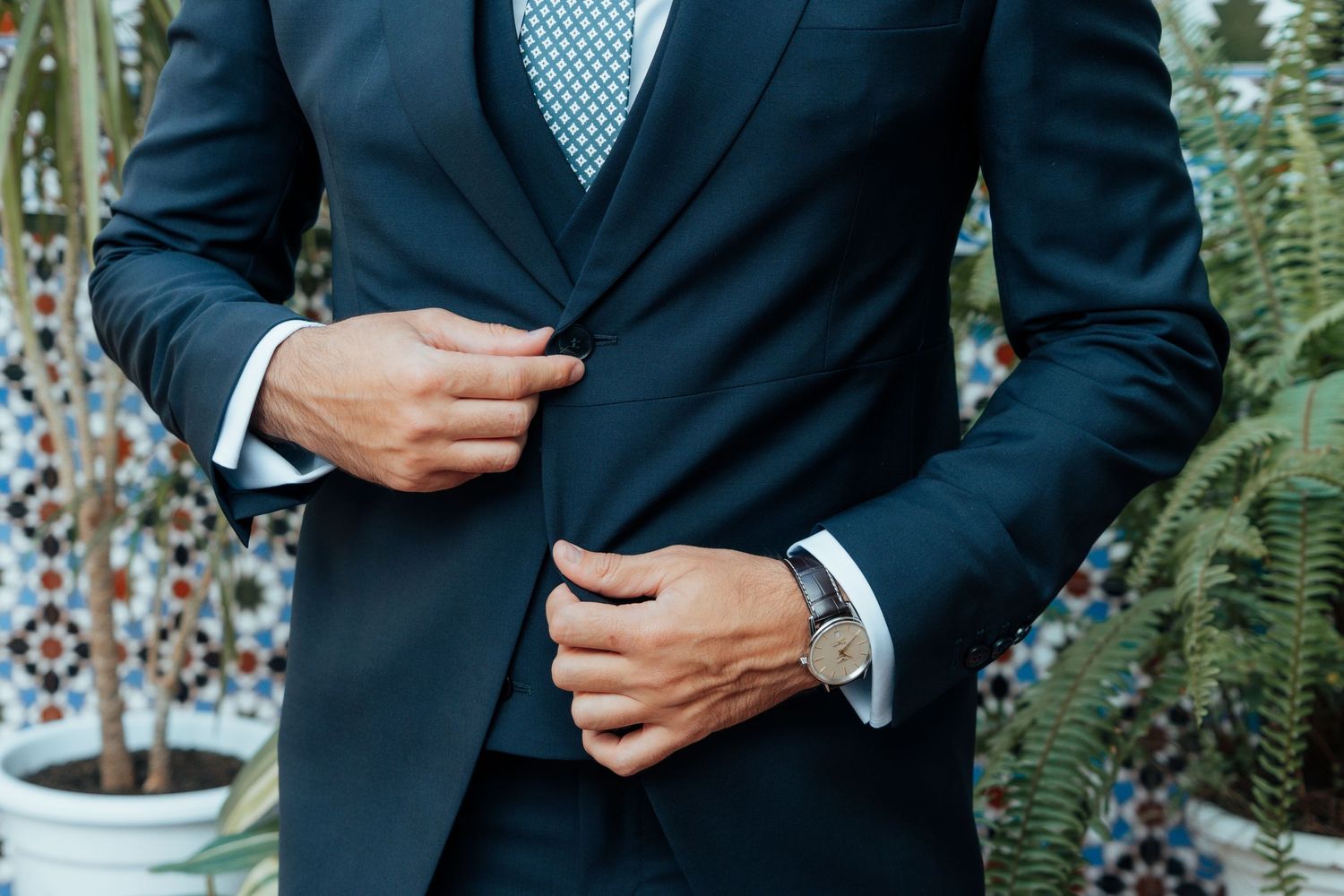 Close-up of groom in navy suit adjusting jacket with silver watch and blue patterned tie.
