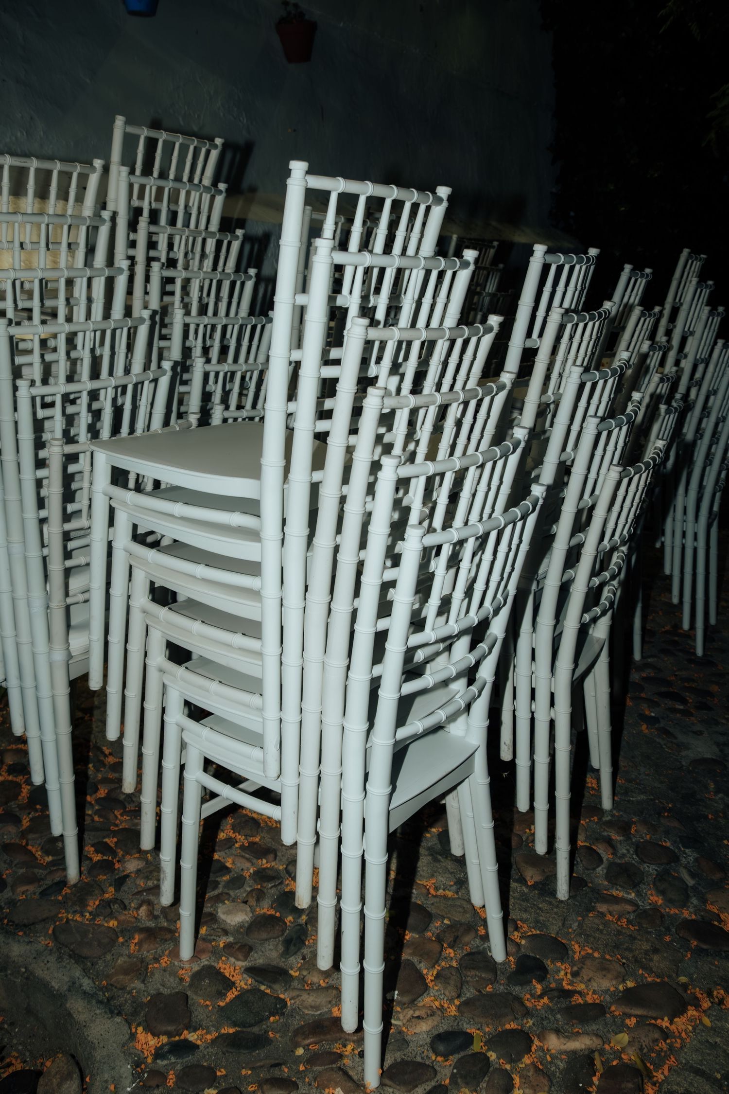 Stacks of white chiavari chairs piled on cobblestone floor after an outdoor evening wedding event.