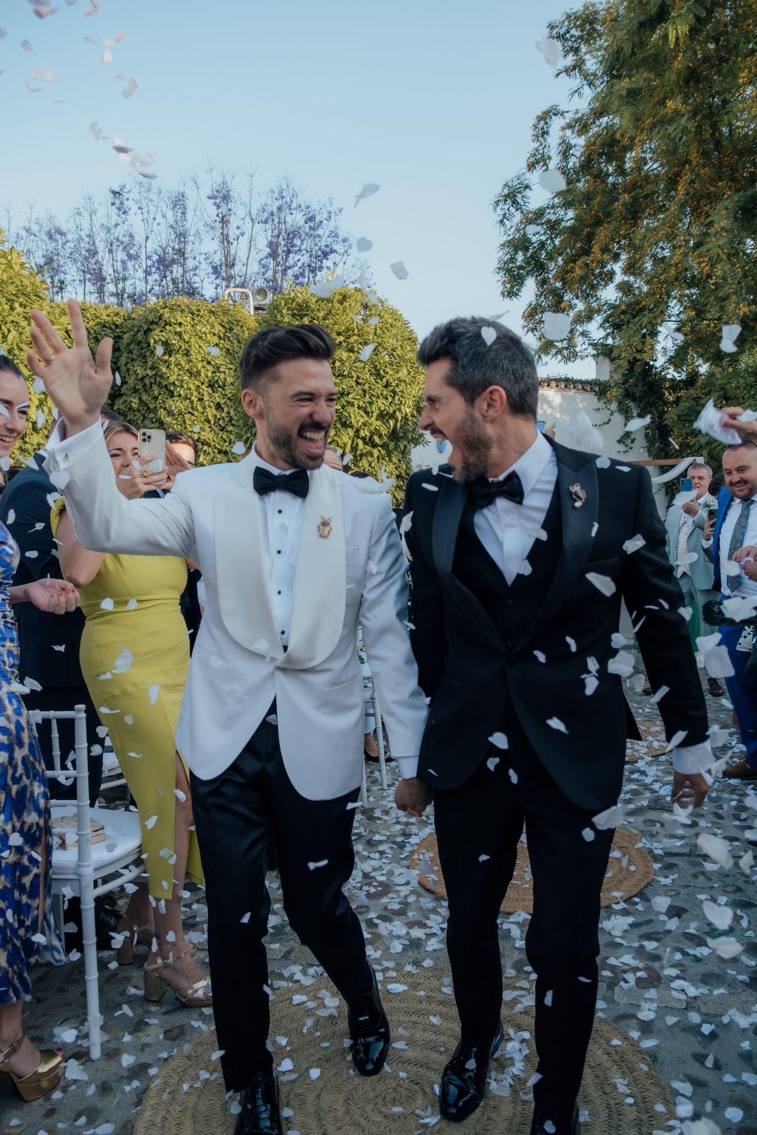 Two grooms laugh together walking through white confetti shower after outdoor wedding ceremony.