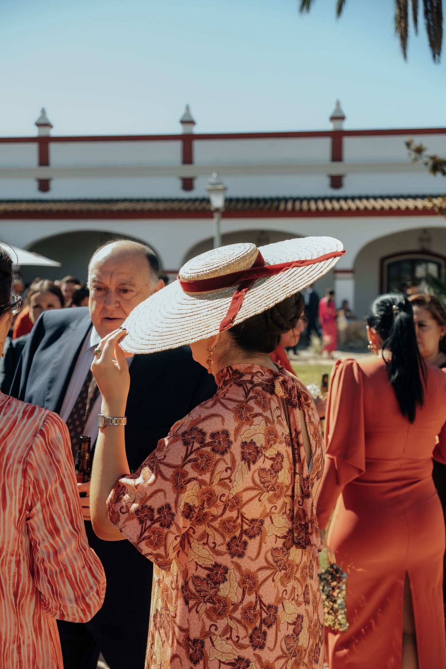 Wedding guest in floral dress and wide-brim straw hat socializes at sunny outdoor Spanish reception.