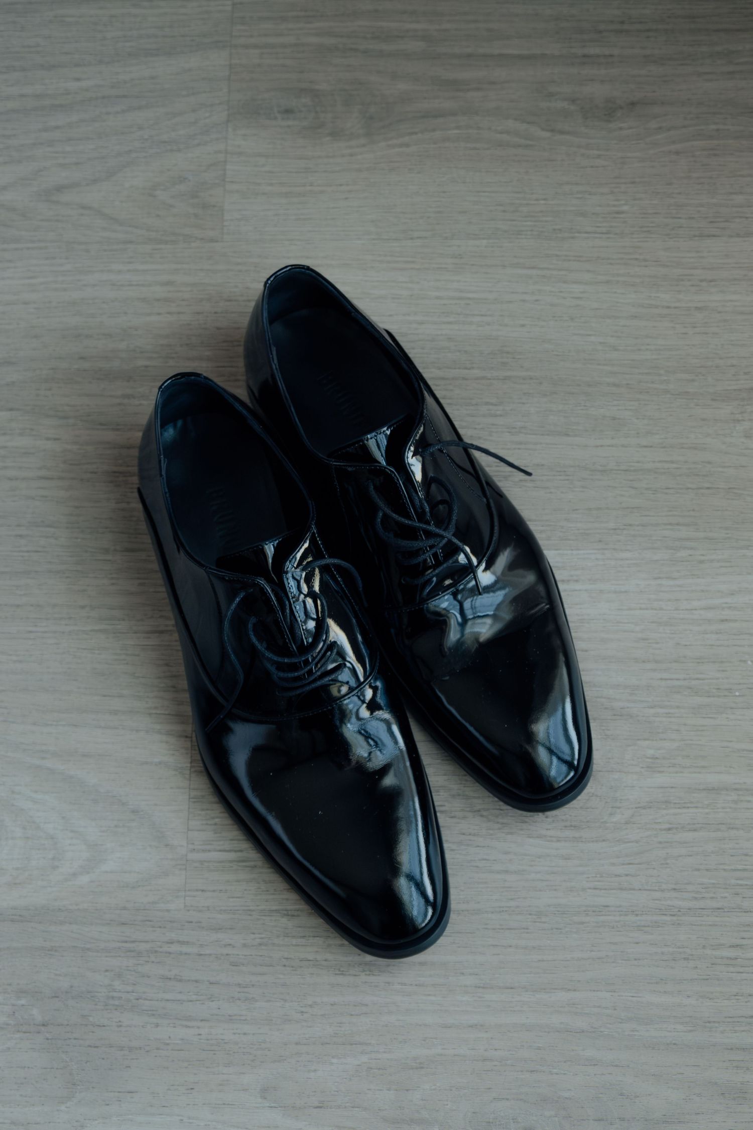 Pair of shiny black patent leather Oxford dress shoes on a wooden floor.