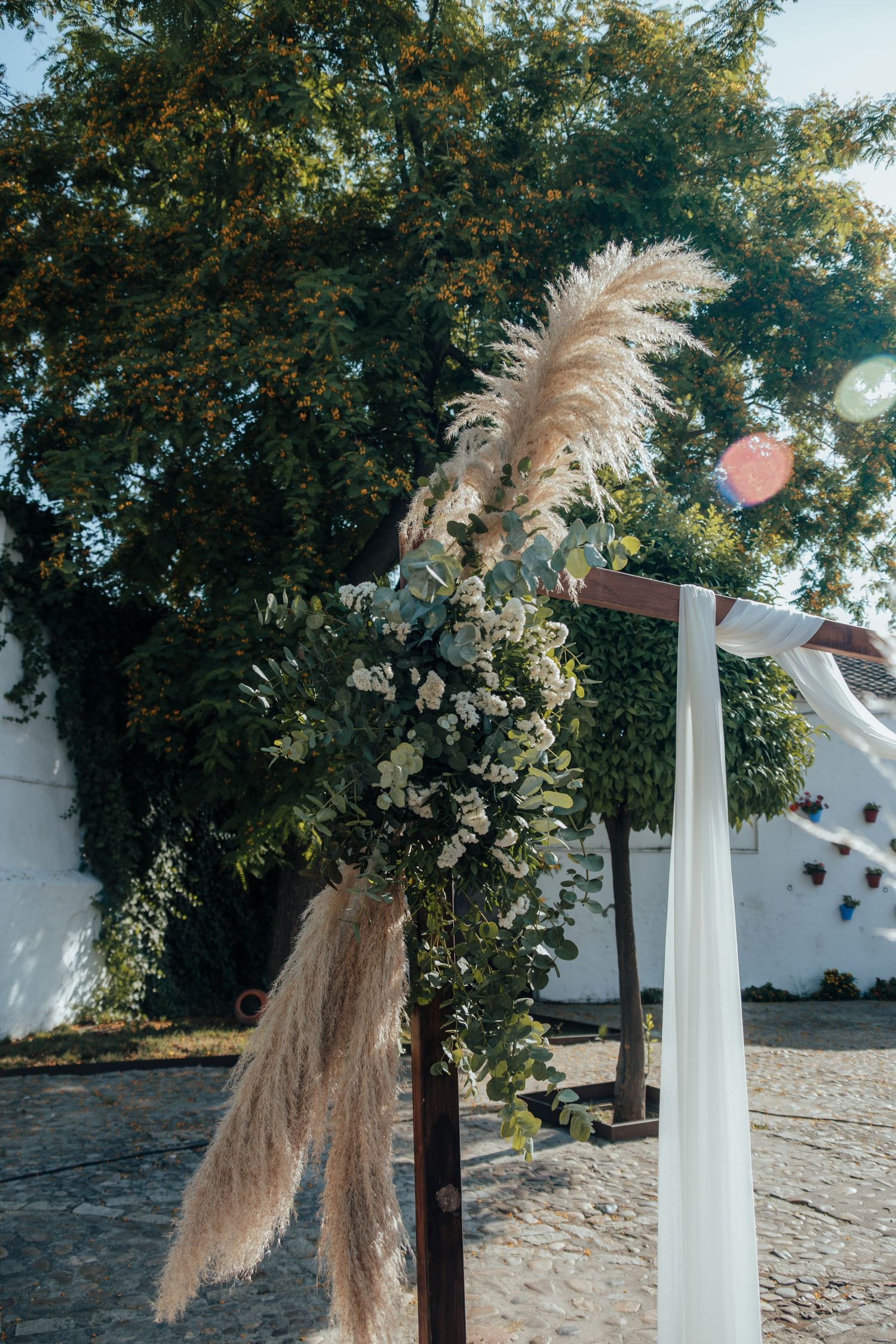 Rustic wooden wedding arch adorned with pampas grass and white flowers against a sunny outdoor backdrop.