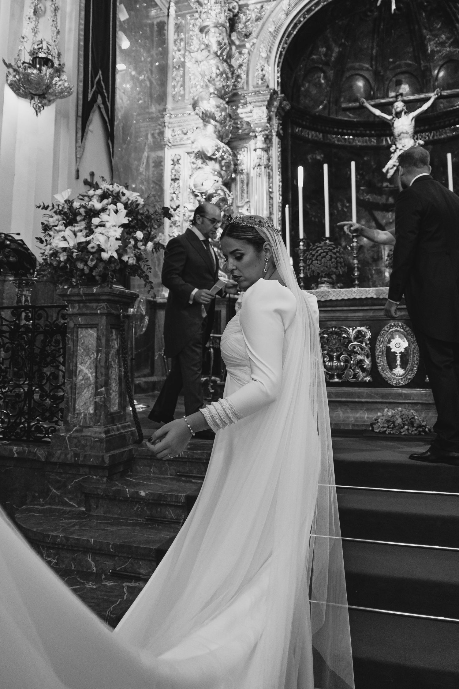 Bride in elegant white gown and veil stands inside ornate church during wedding ceremony.