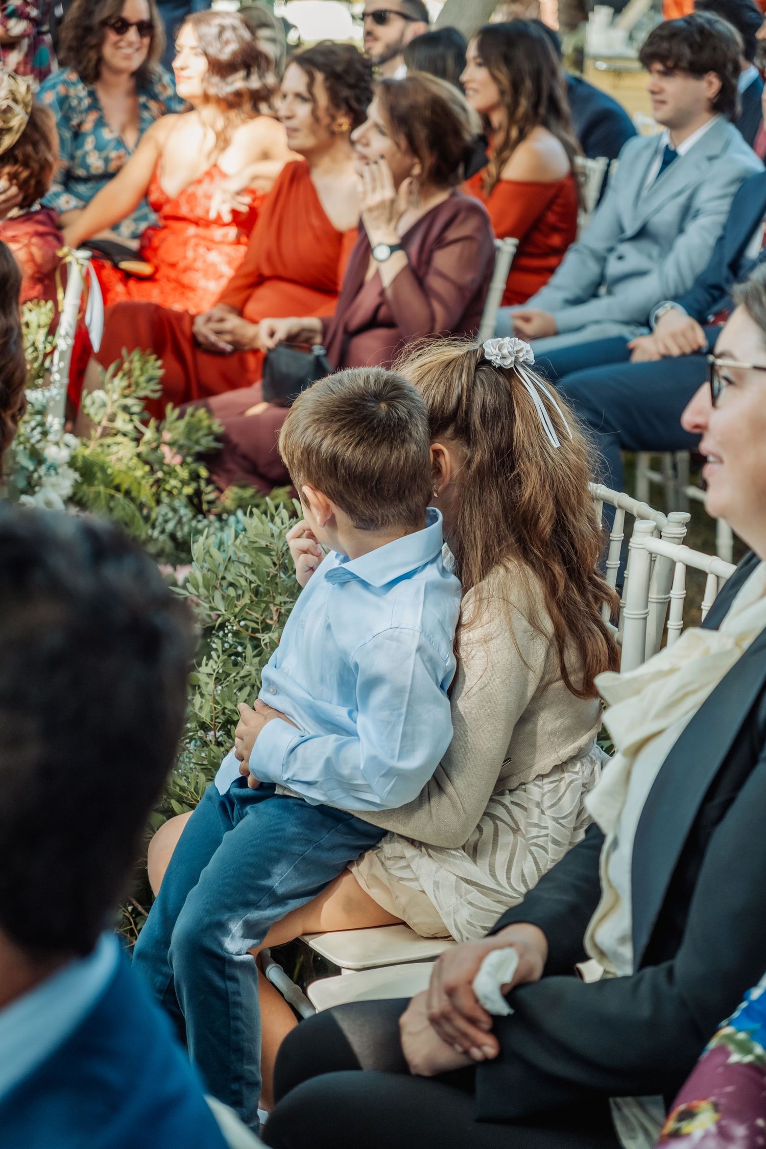 Young boy sitting on woman's lap during outdoor wedding ceremony with colorful guests seated in white chairs.