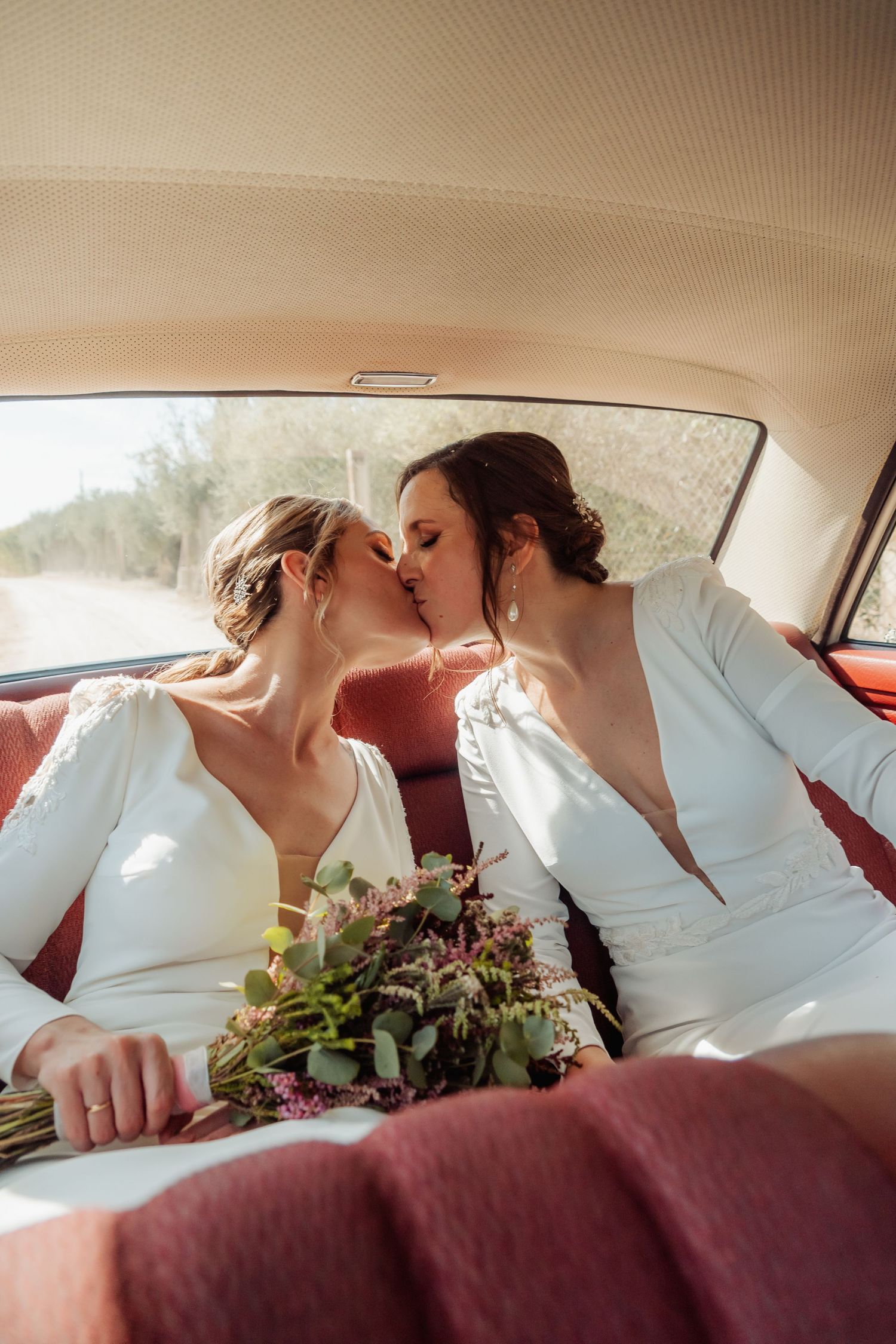Two brides share a romantic kiss in the backseat of a vintage car holding a floral bouquet.