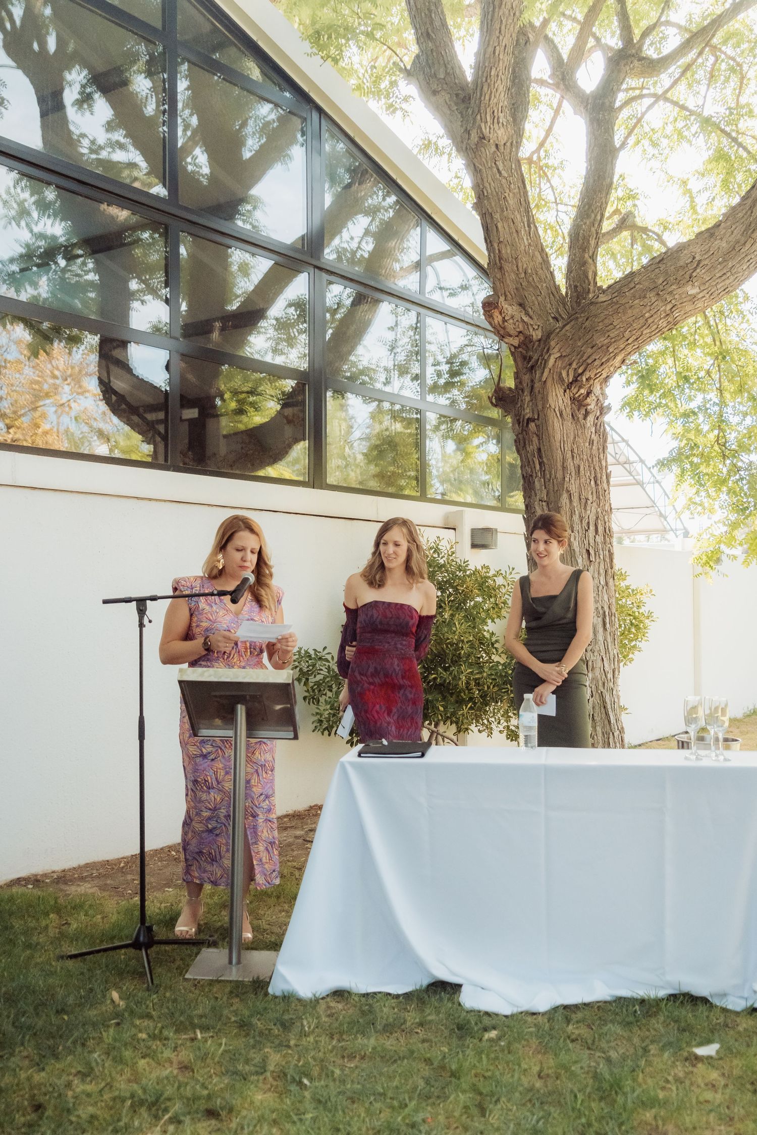 Three women stand at a podium with microphone during an outdoor wedding reception speech near a large tree.