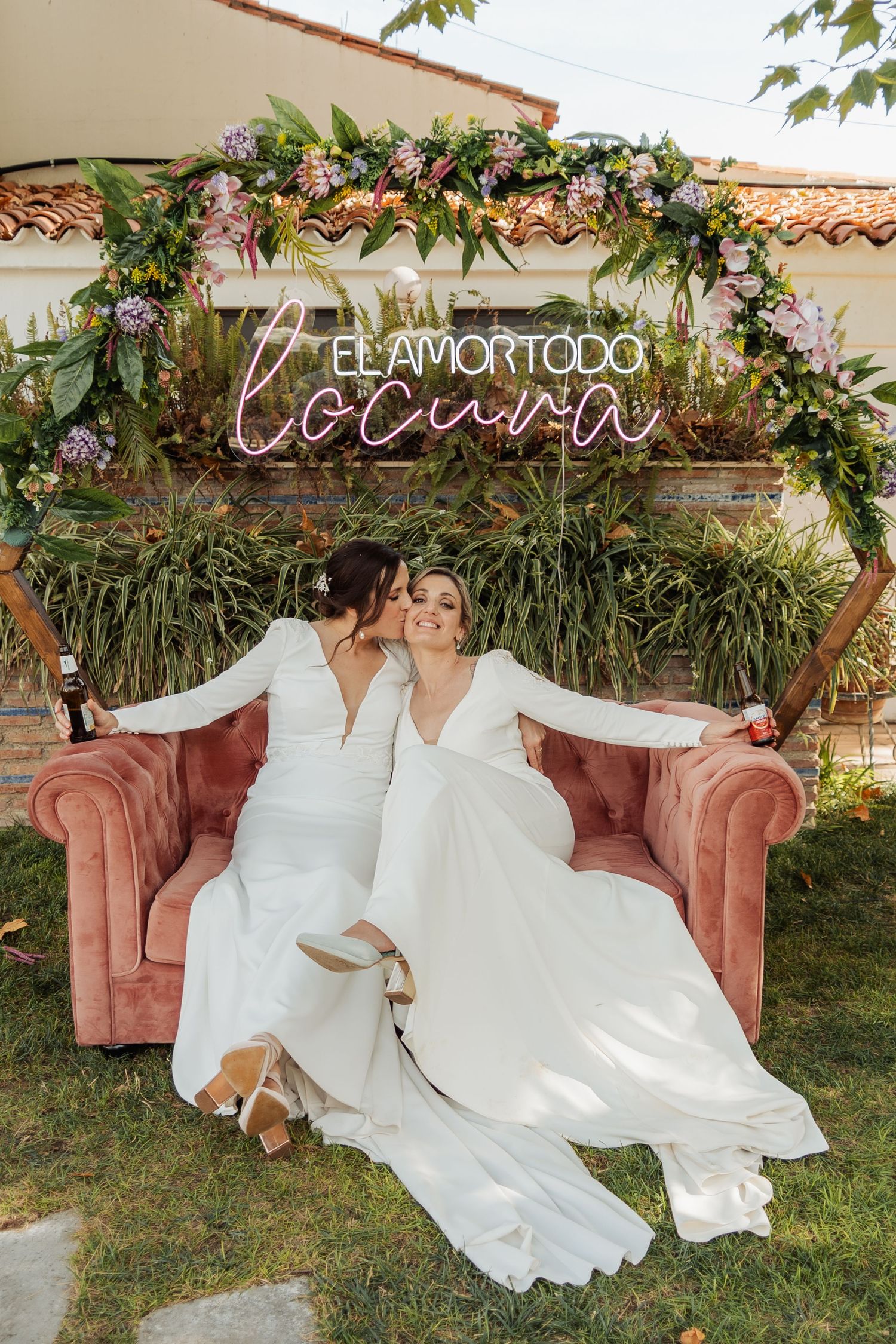 Two brides in white gowns sit on a pink velvet sofa under a floral arch with neon sign at wedding.