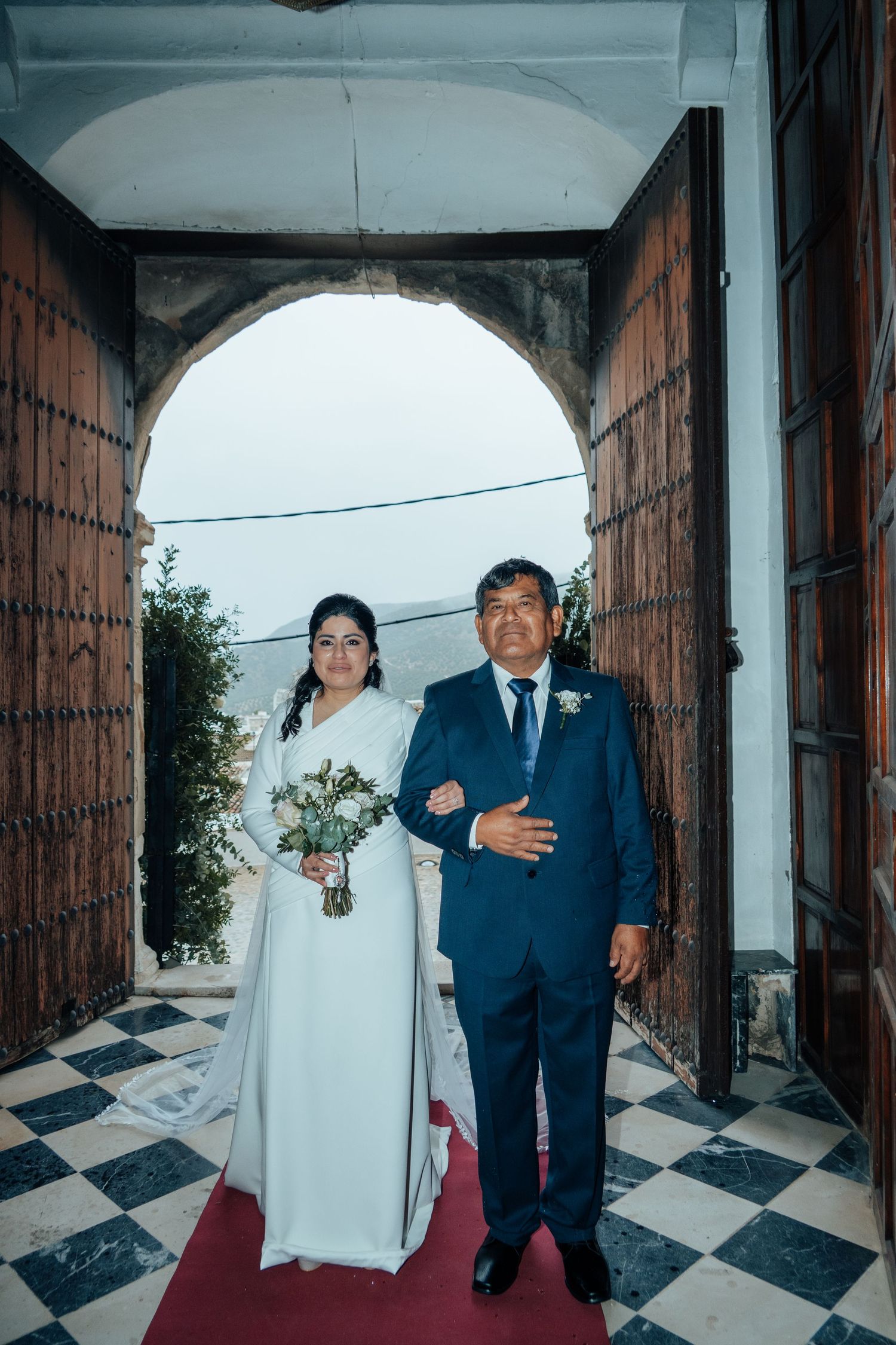 Bride in white gown holding bouquet walks with father through arched church entrance on red carpet.