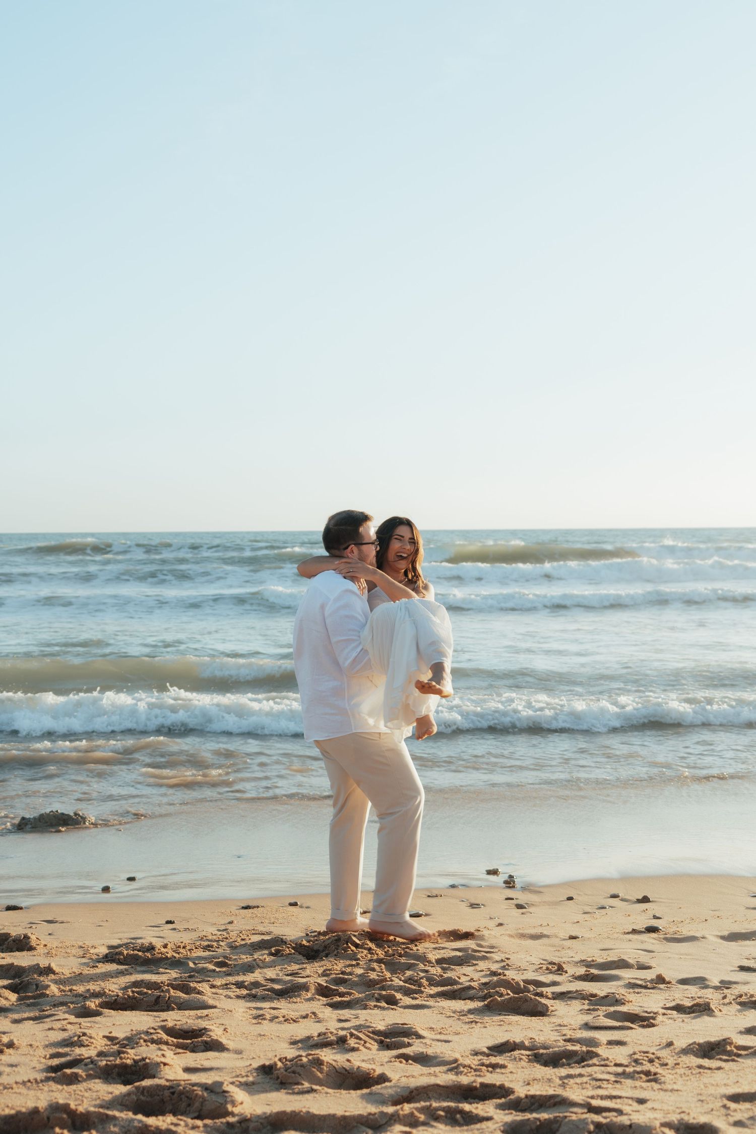 Romantic couple embracing on a sandy beach with ocean waves, both wearing white outfits in warm sunlight.