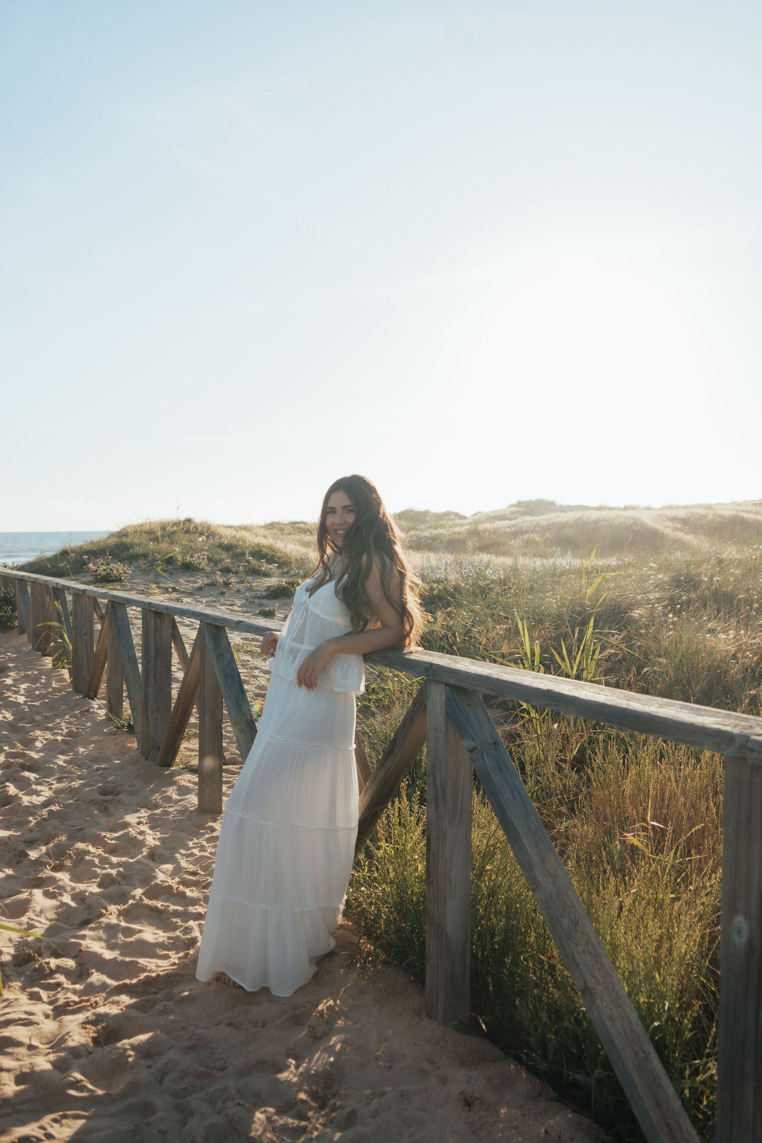 Woman in white maxi dress leaning on wooden beach railing with sandy dunes and ocean in background.