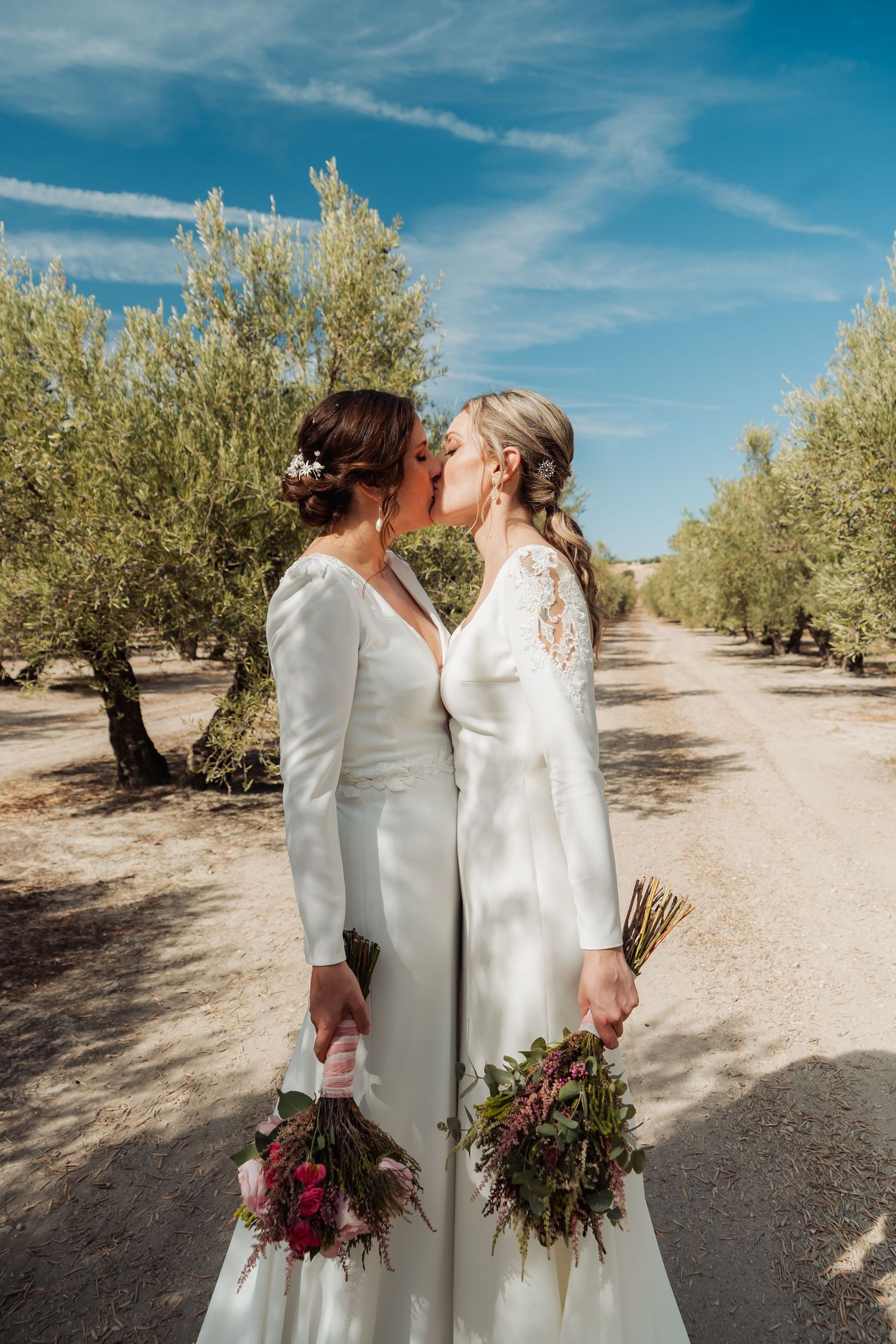 Two brides in white gowns kissing while holding bouquets on a tree-lined path under a blue sky.