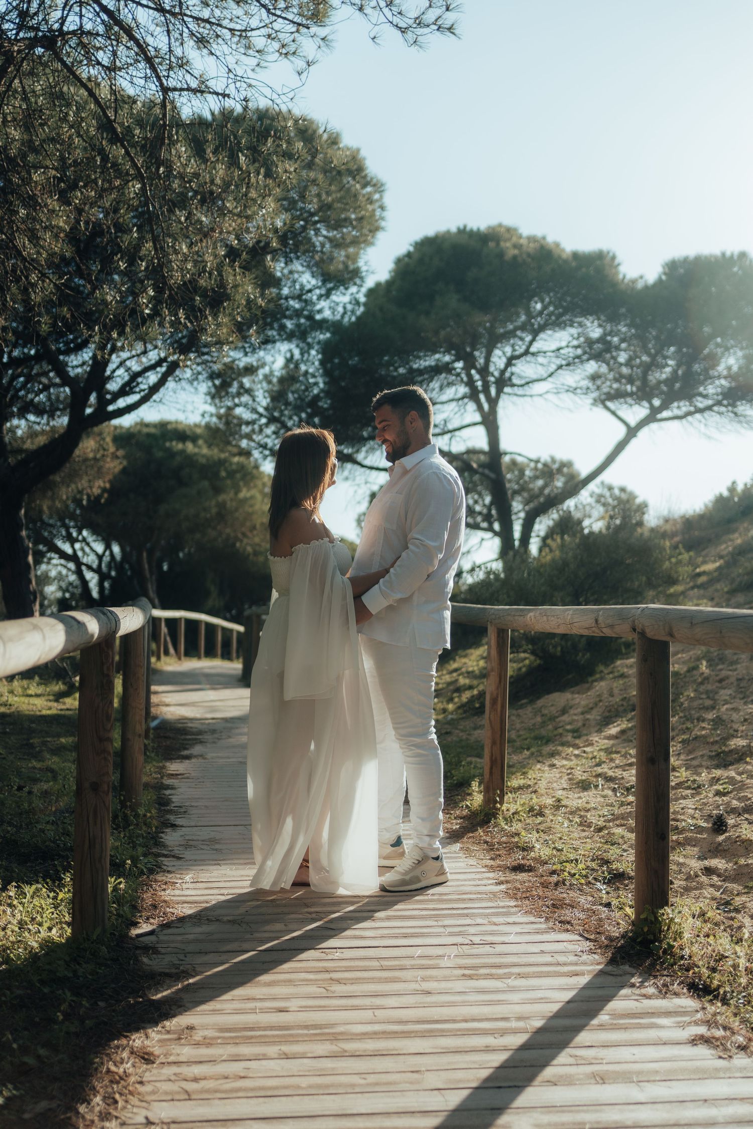 Couple in white outfits embracing on a wooden boardwalk surrounded by trees in warm sunlight.