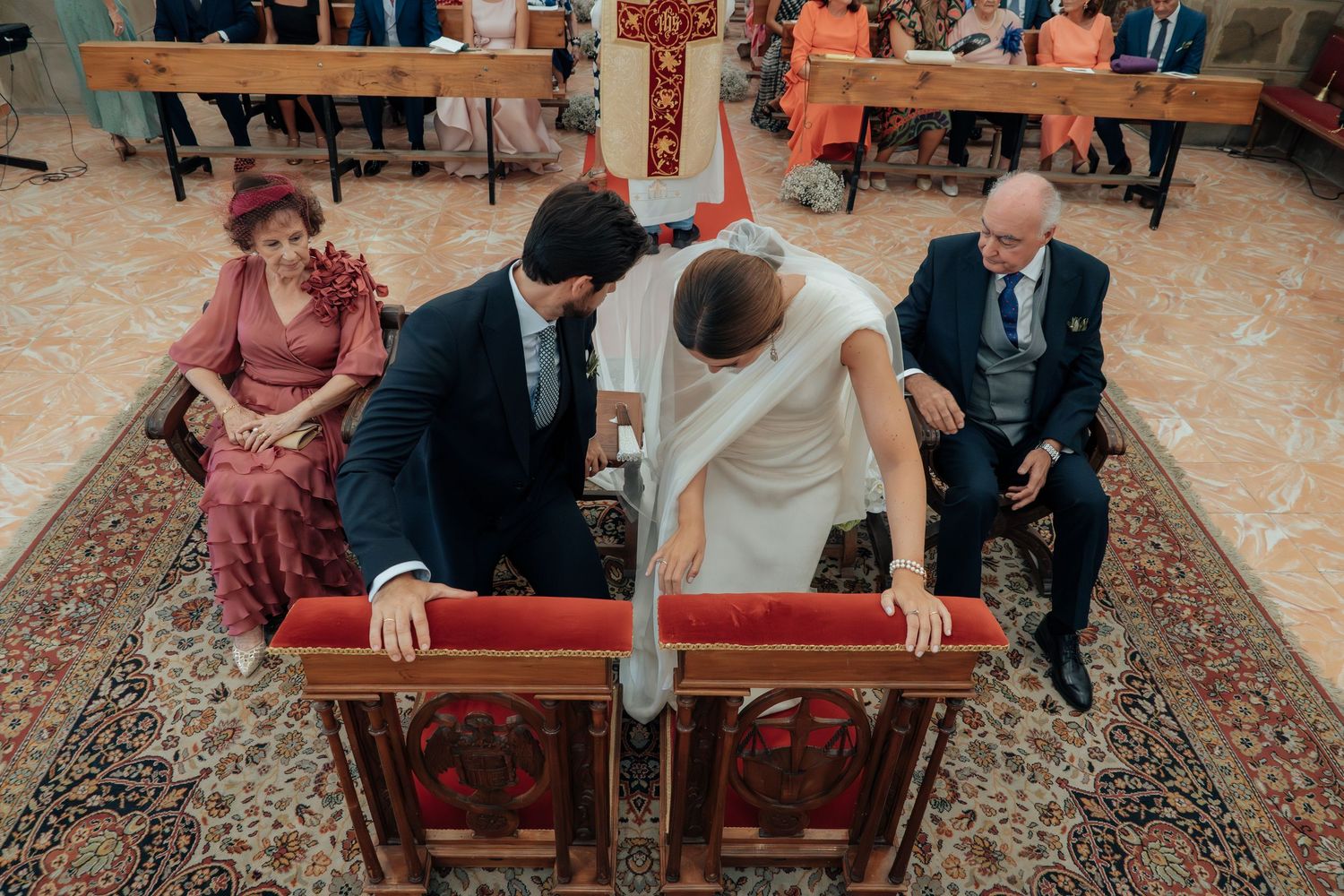 Overhead view of bride and groom kneeling at church altar during wedding ceremony with seated family members.