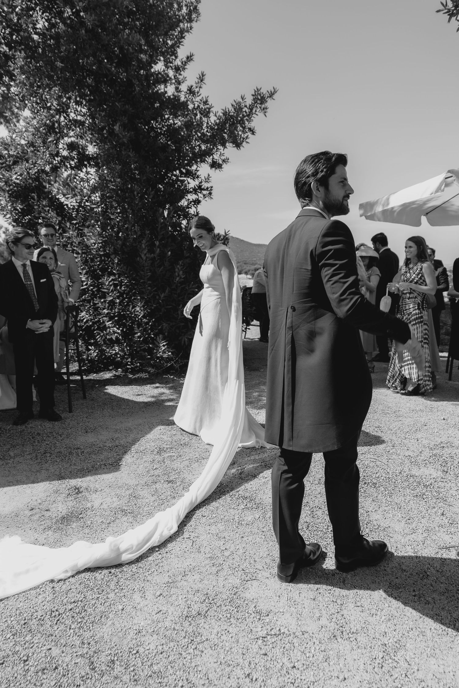 Black and white photo of bride walking toward groom during outdoor wedding ceremony with guests in background.
