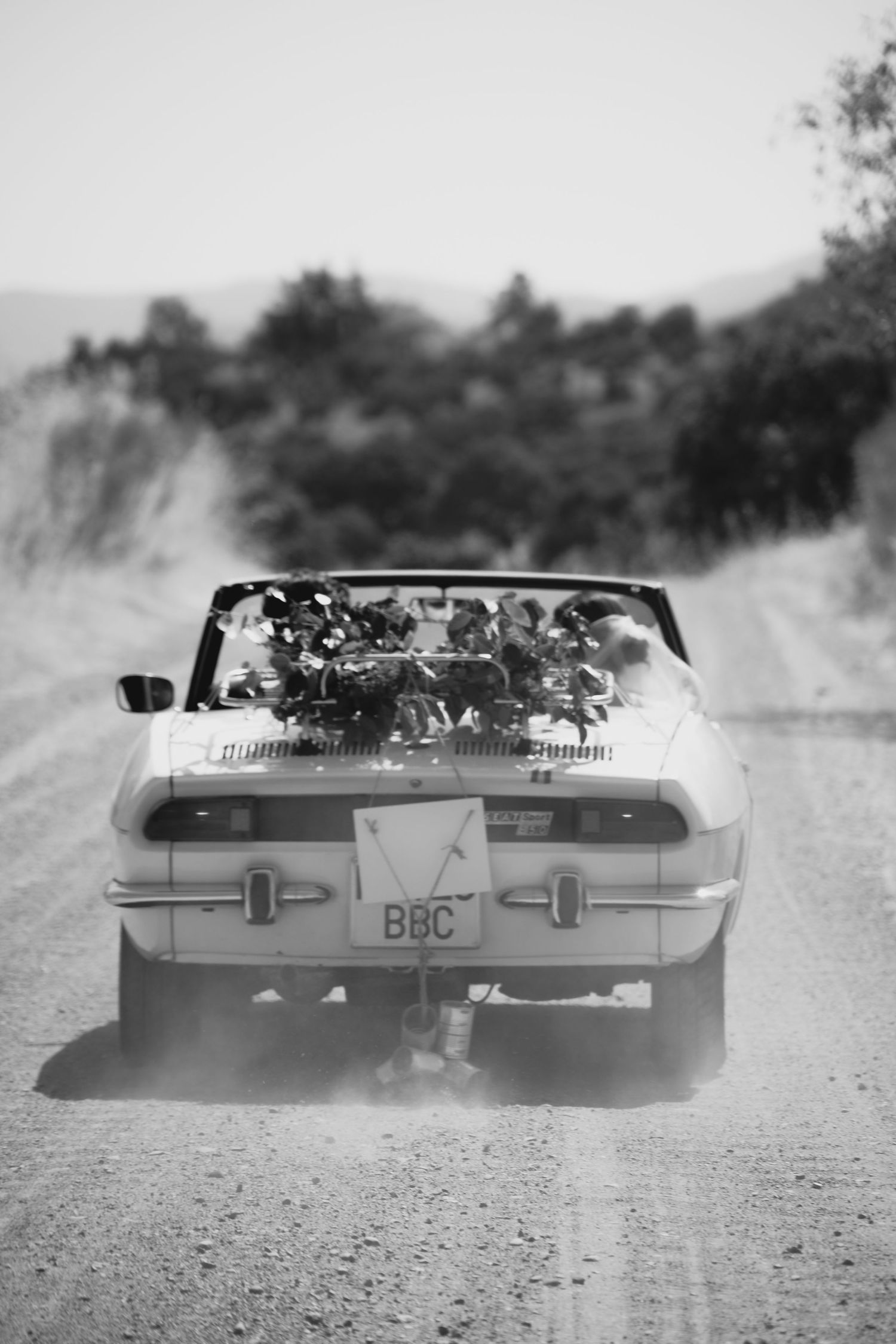 Black and white photo of newlyweds driving away in a white convertible on a dusty rural road.