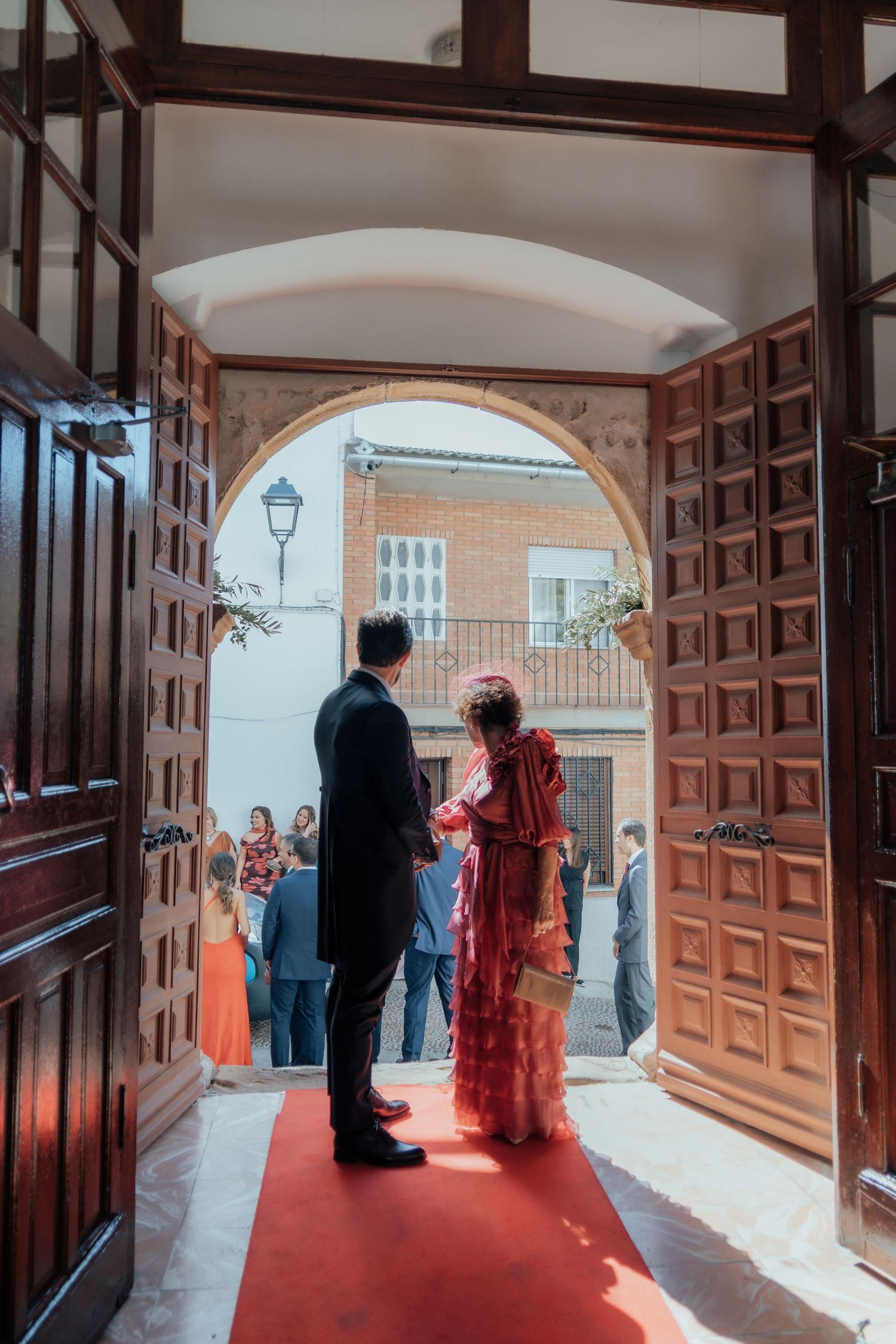 Groom in black suit and bride in red dress stand at church entrance on red carpet.