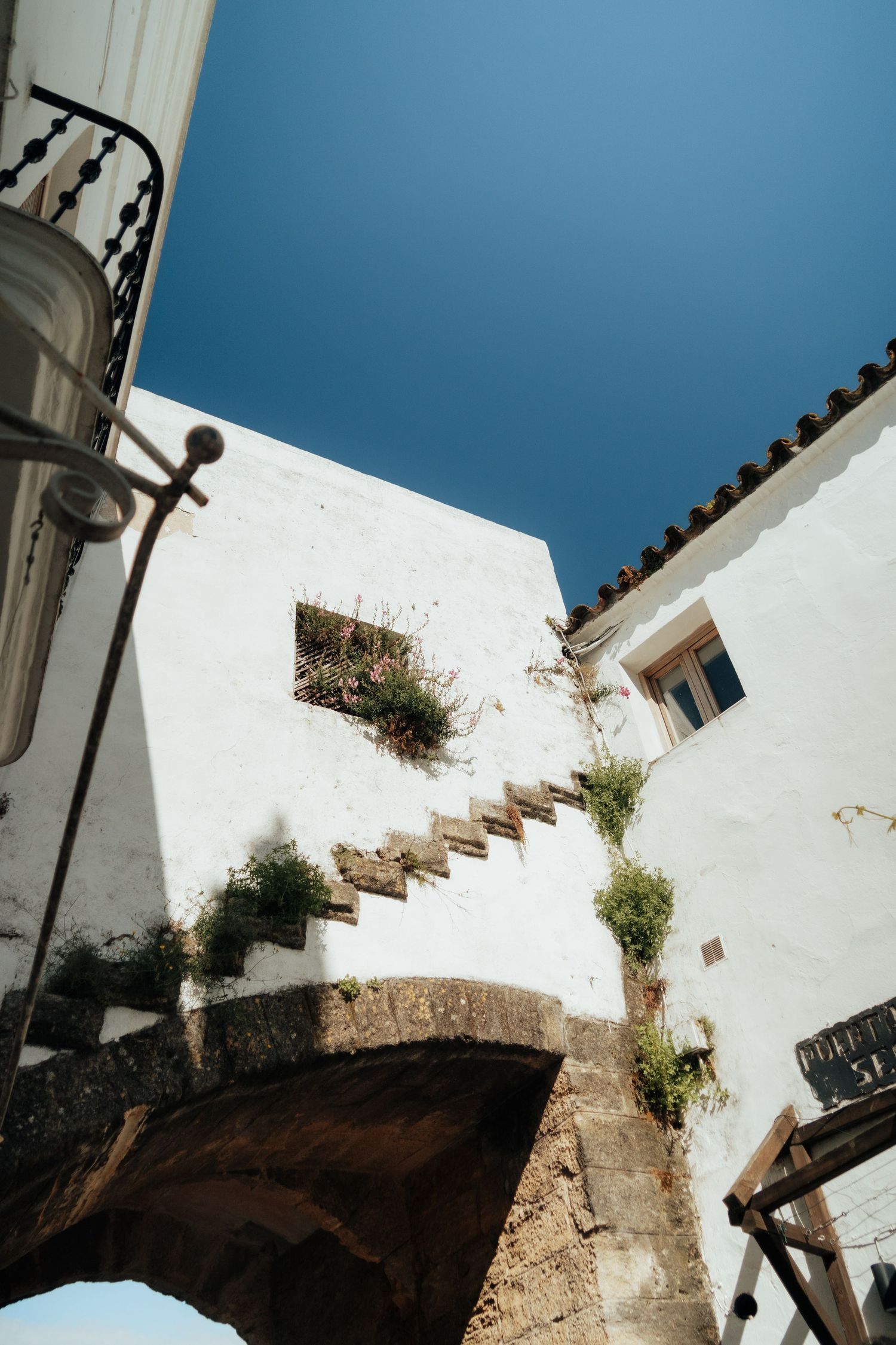 White-washed Mediterranean building with stone staircase, climbing plants, and wrought iron balcony under deep blue sky.