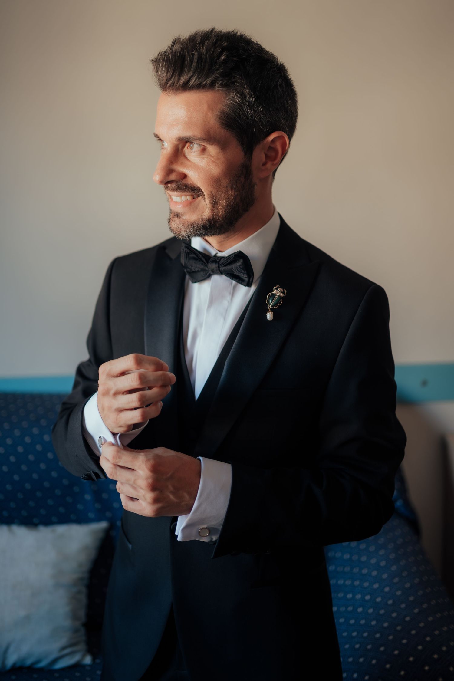 A handsome bearded groom in a black tuxedo with bow tie and boutonniere adjusts his cufflinks.