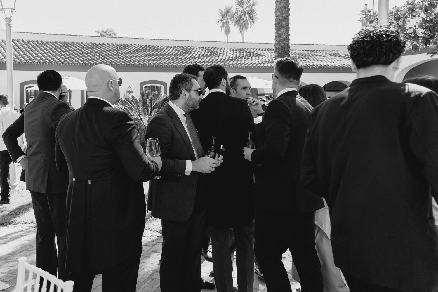 Group of men in suits socializing outdoors at wedding reception in black and white.