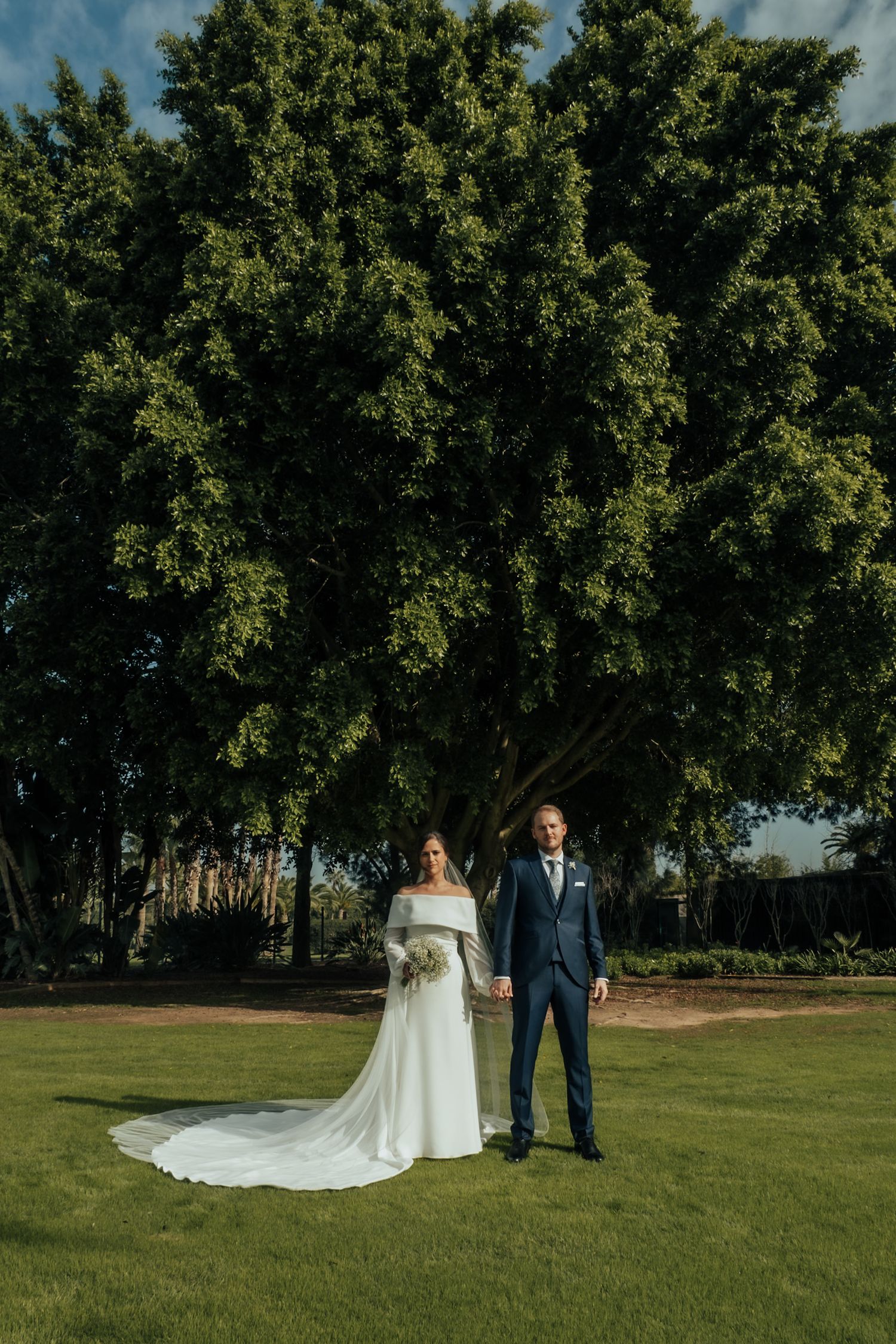 Bride in off-shoulder white gown with long train and groom in navy suit pose before a large green tree.