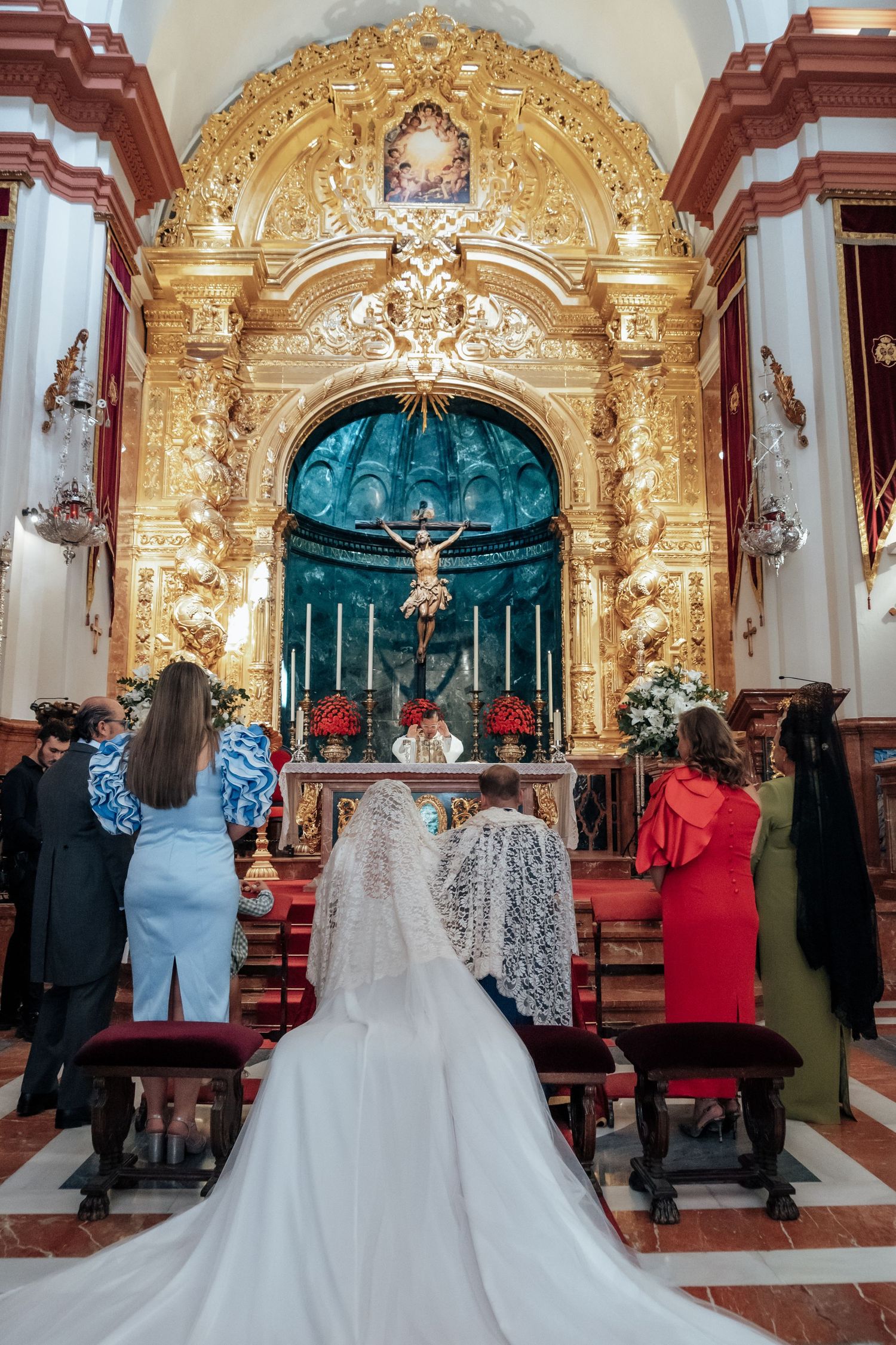 Bride in lace mantilla veil praying at ornate golden baroque church altar during wedding ceremony.