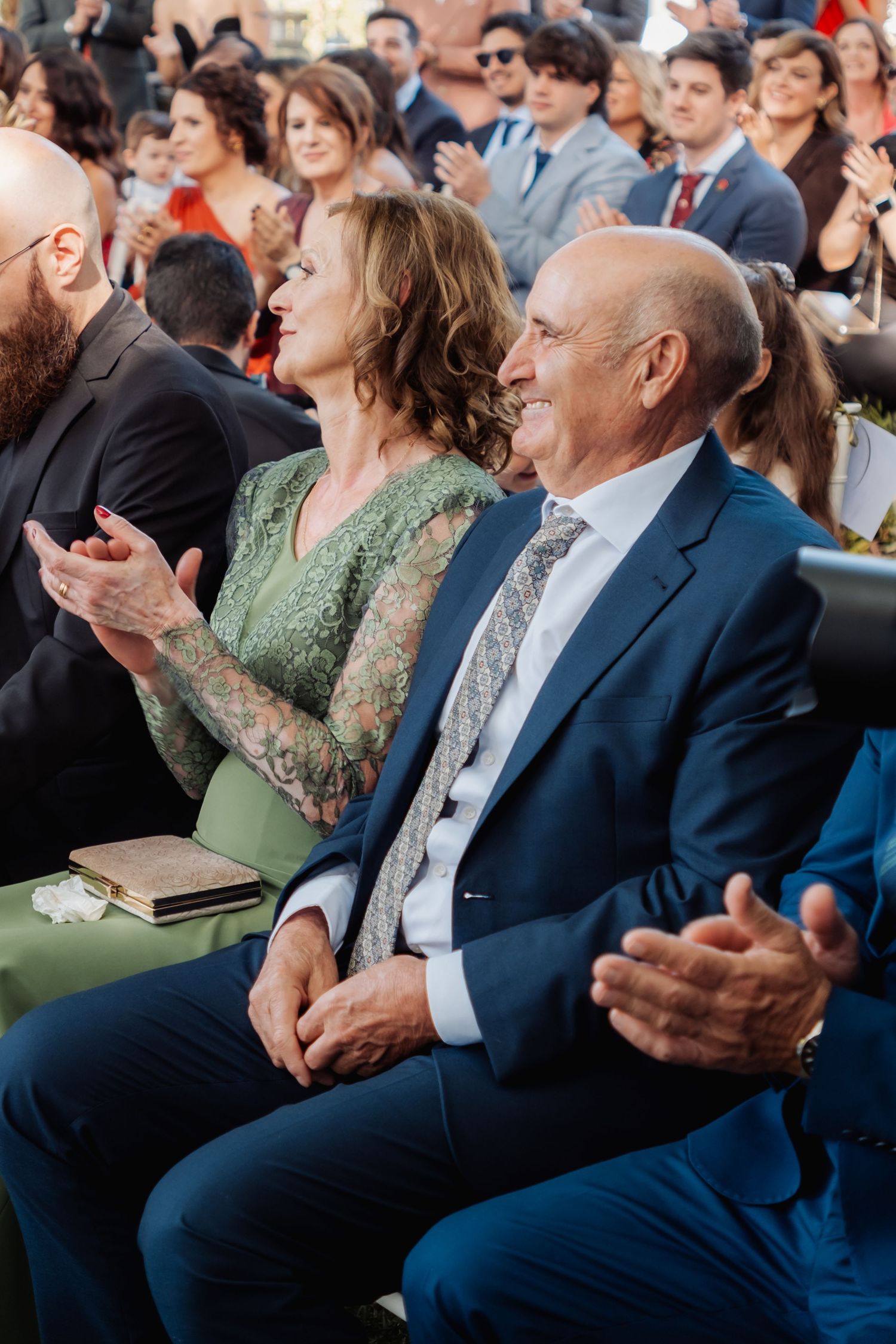 Wedding guests in formal attire clapping and smiling during outdoor ceremony celebration.