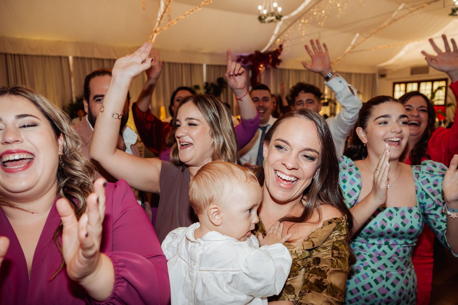 Joyful wedding guests celebrating on dance floor with baby in arms and raised hands.