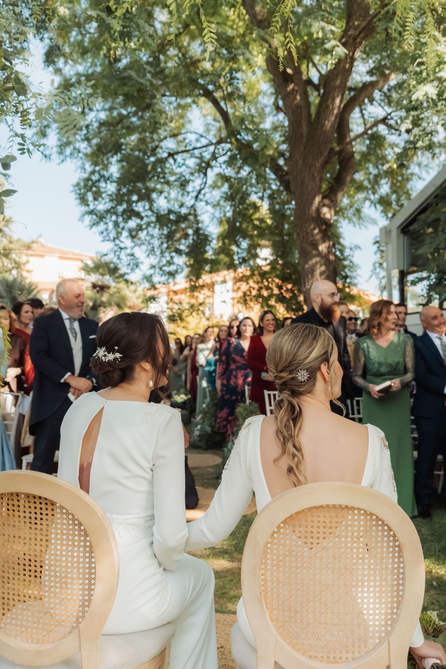 Two brides in white gowns sit in rattan chairs during outdoor garden wedding ceremony.
