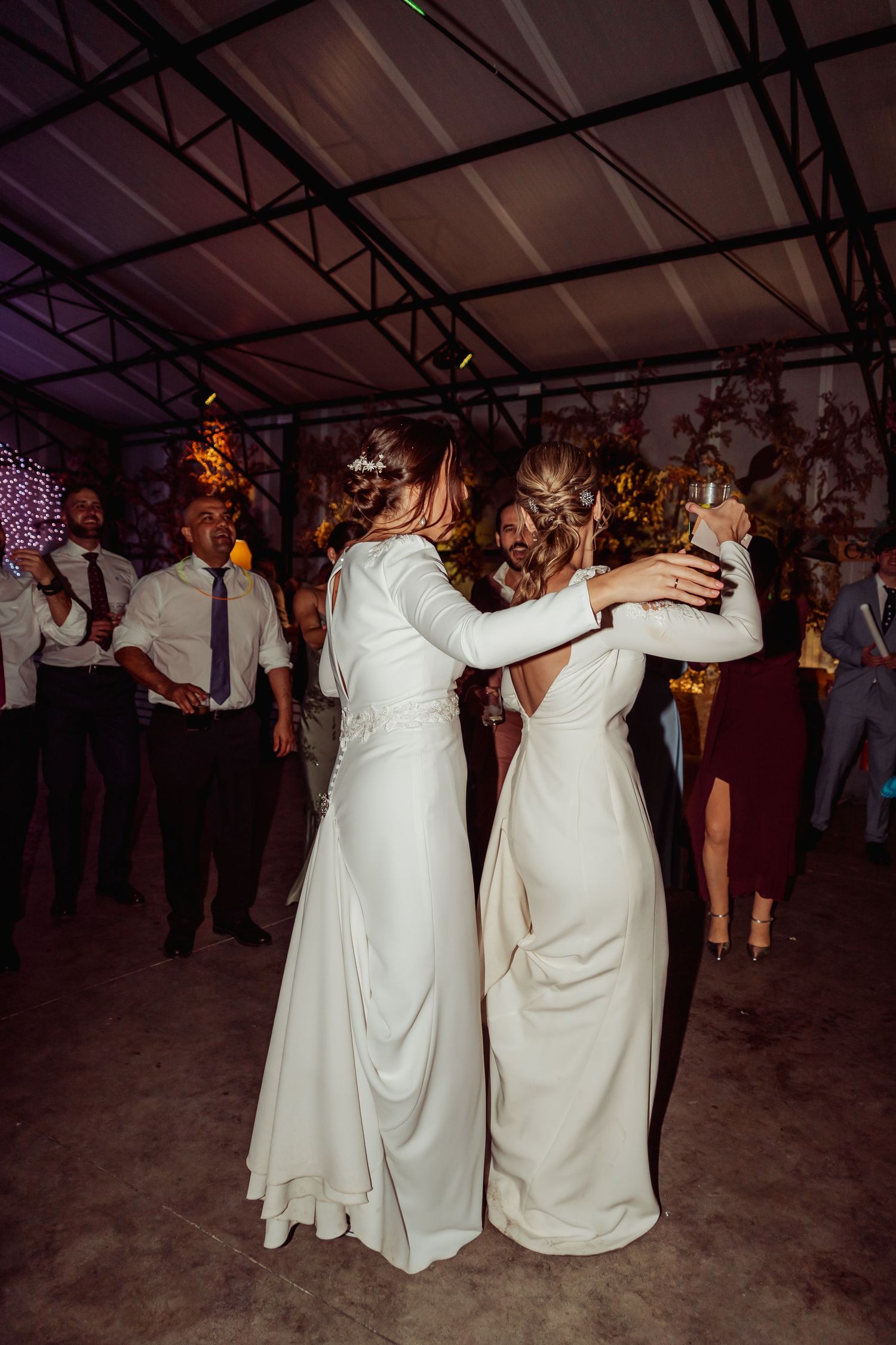 Two brides in white wedding gowns sharing a first dance embrace surrounded by wedding guests.